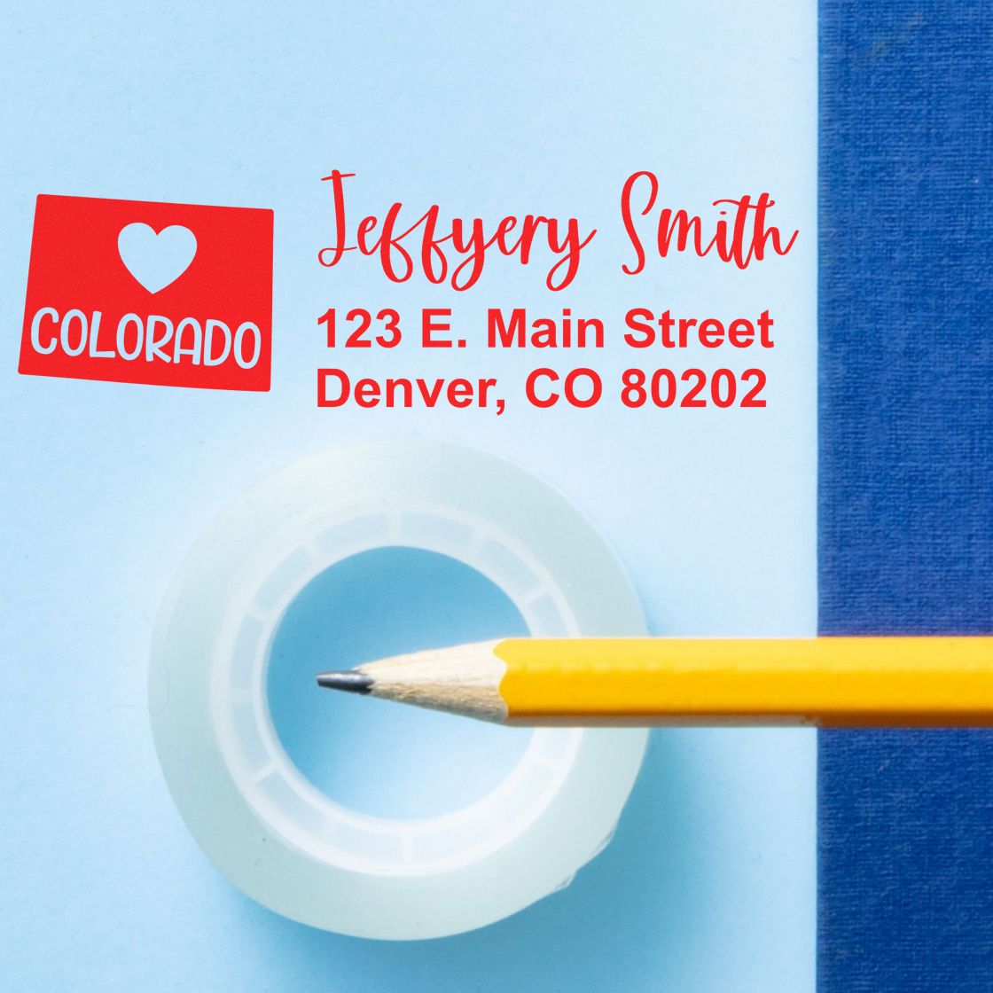 Slim Colorado Custom Address Stamp for Envelopes displayed on a blue surface with a pencil and tape, featuring a red heart design and sample address: Jeffery Smith, 123 E. Main Street, Denver, CO 80202.