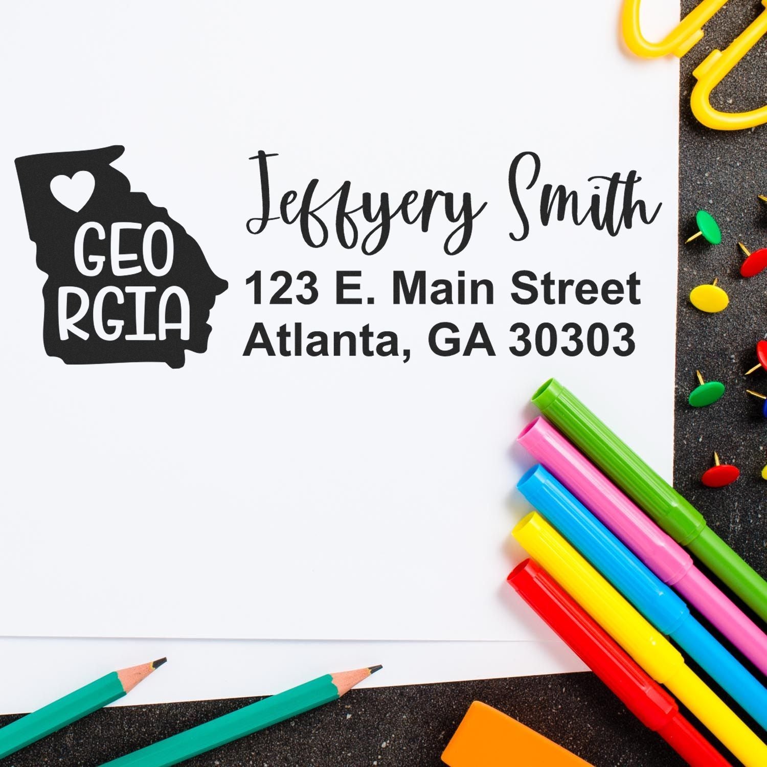 Slim Georgia Custom Address Stamp for Envelopes on white paper with colorful pens and paperclips. Features a heart-shaped Georgia design and personalized address for Jeffery Smith in Atlanta, GA.