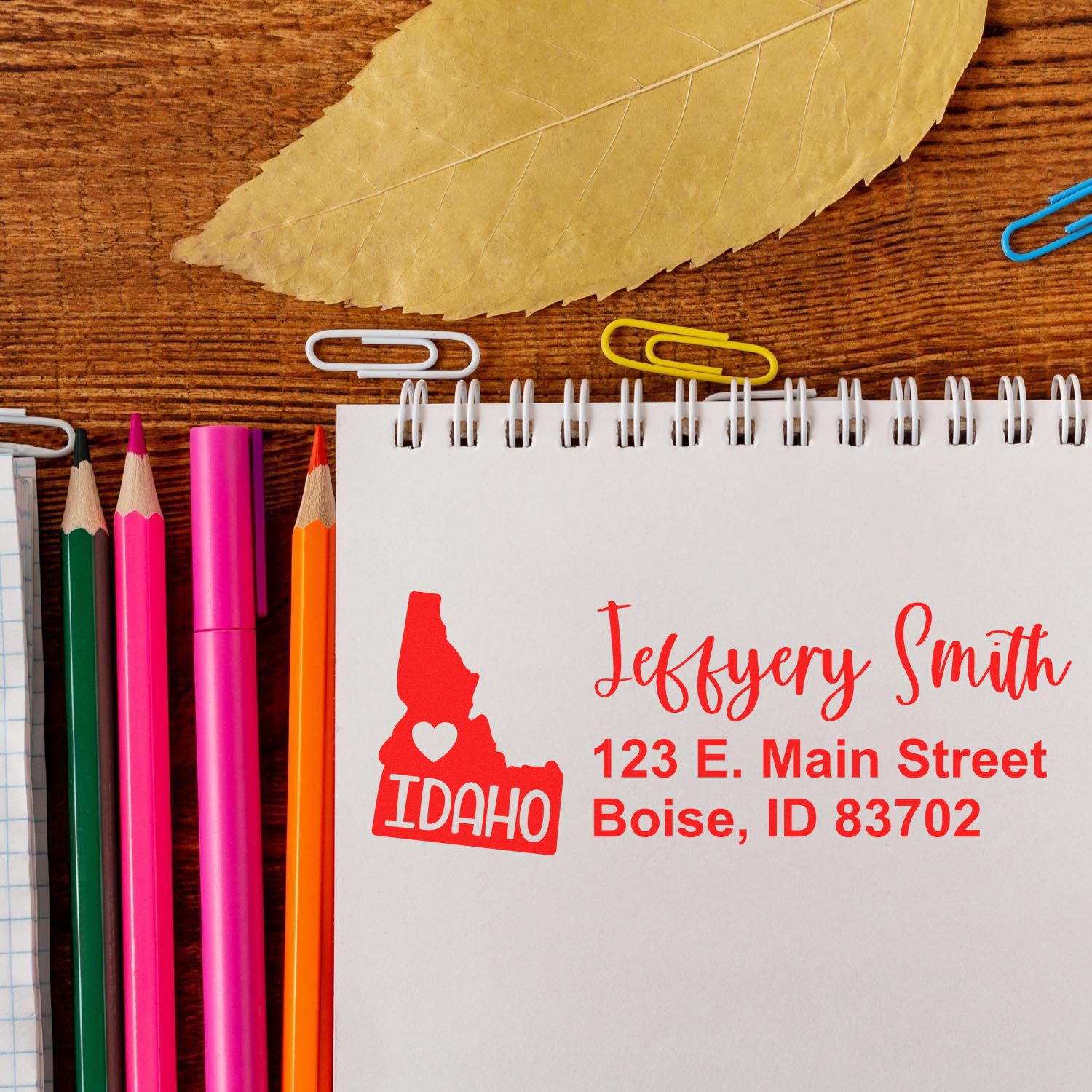 Slim Idaho Custom Address Stamp for Envelopes displayed on a notepad with colorful pencils and paperclips, featuring a red Idaho state outline and personalized address in bold red text.