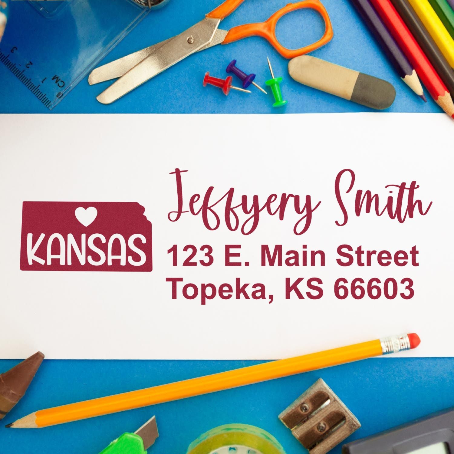 Kansas State Love Personalized Address Stamp on white paper with Jeffery Smith, 123 E. Main Street, Topeka, KS 66603 in red. Surrounded by colorful office supplies on a blue background.