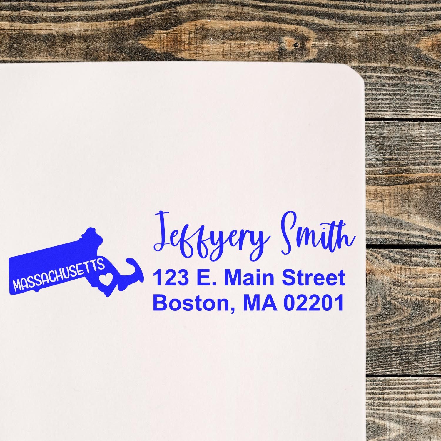 Slim Massachusetts Custom Address Stamp for Envelopes on white paper, featuring a blue state outline with a heart and personalized address in elegant font. Placed on a rustic wooden surface.