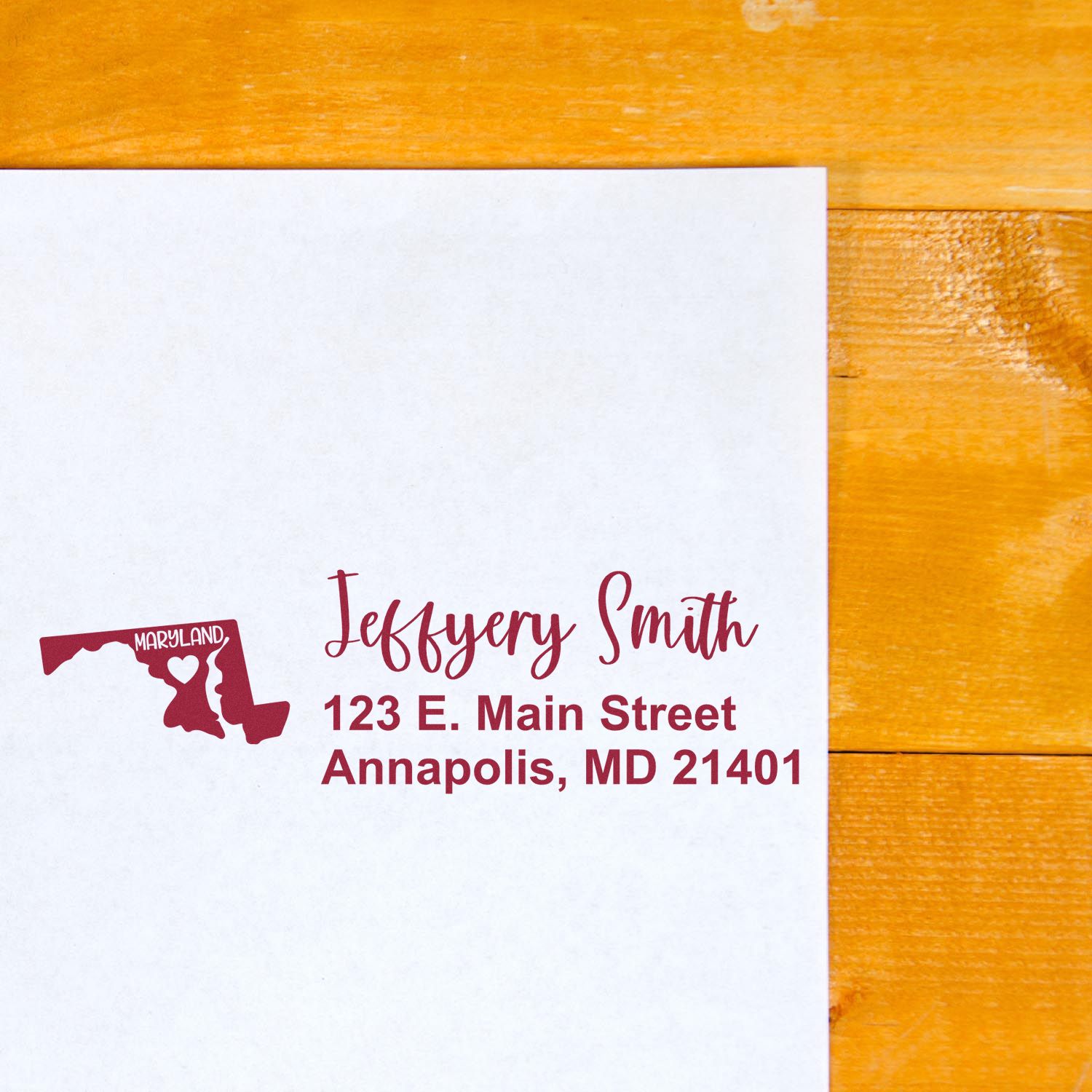 Slim Maryland Custom Address Stamp for Envelopes displayed on a white envelope with a red Maryland state outline and personalized address in elegant script, placed on a wooden surface.