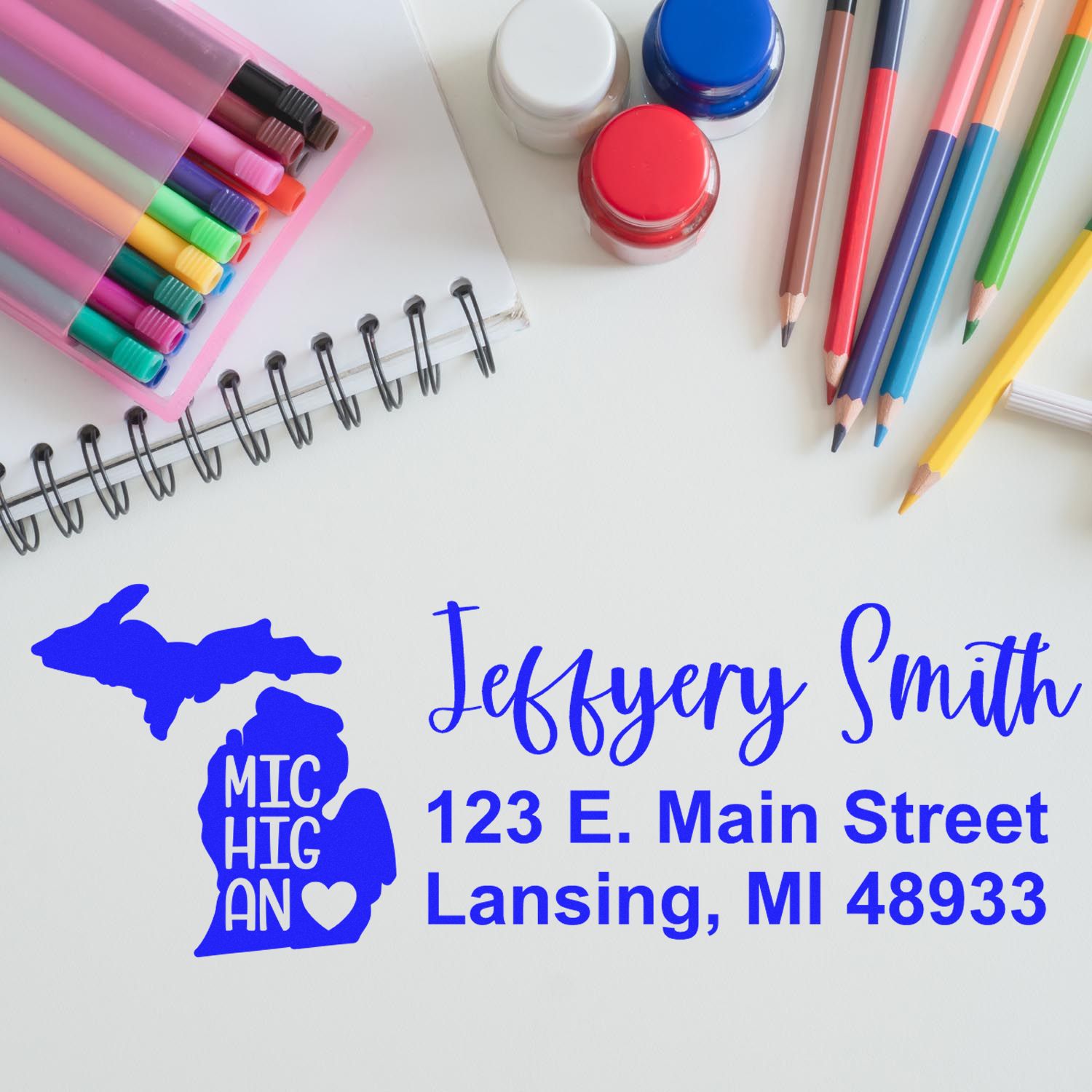 Image of the Michigan State Love Personalized Address Stamp on paper, surrounded by colorful pens, markers, and paint jars. The stamp features a heart design and a sample address in blue ink.