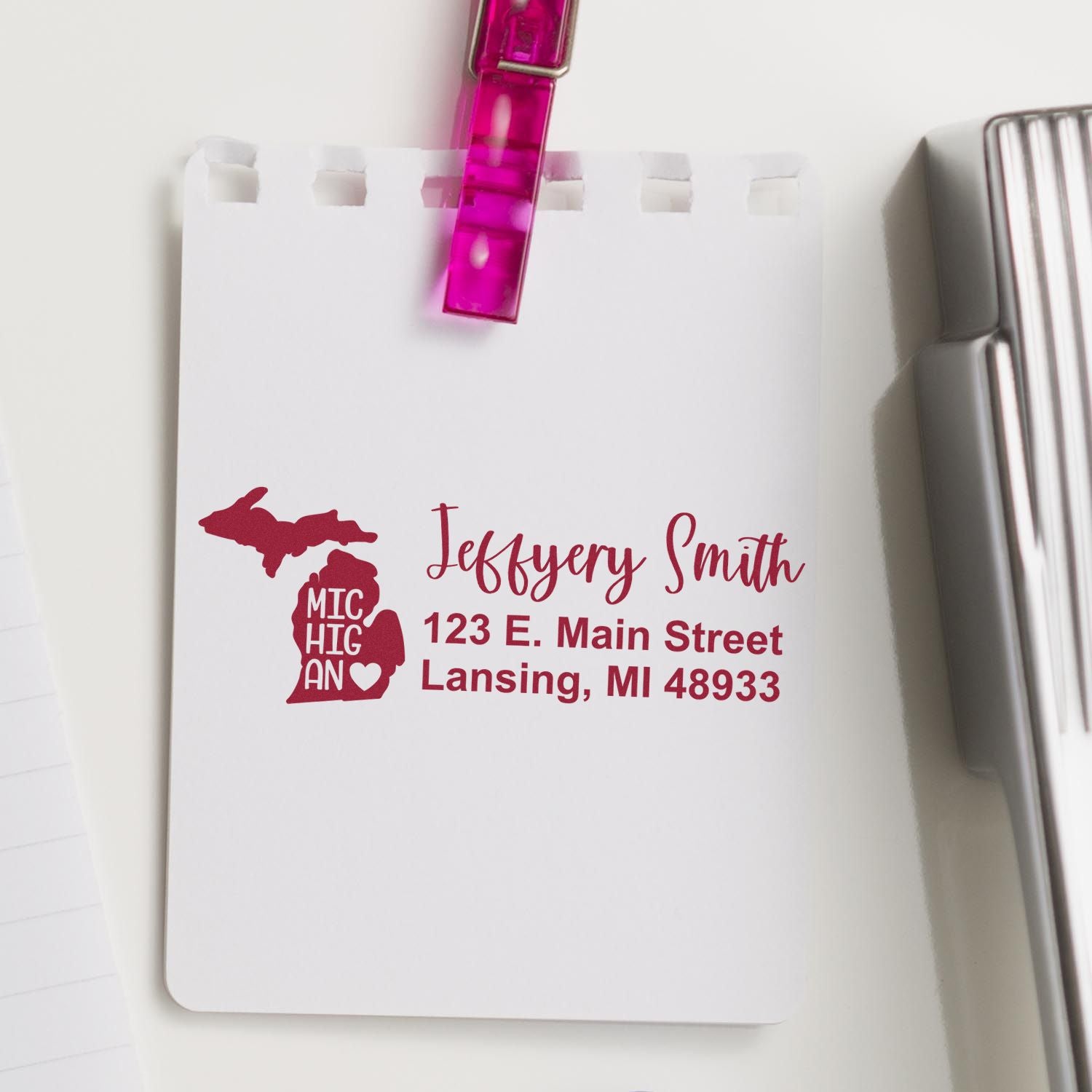 Michigan State Love Personalized Address Stamp on white paper, featuring a red Michigan map design with address details. A pink clip holds the paper, surrounded by office supplies.