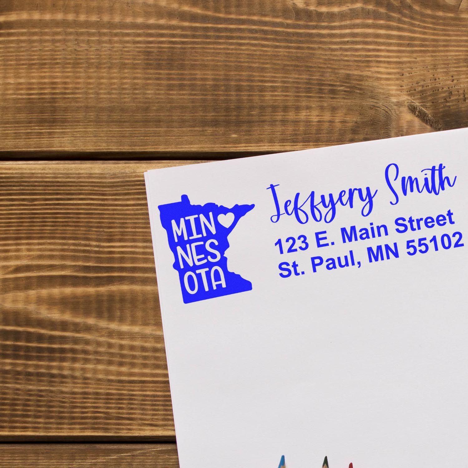 Slim Minnesota Custom Address Stamp for Envelopes displayed on a wooden table, featuring a blue state outline and personalized address in bold font on a white envelope.