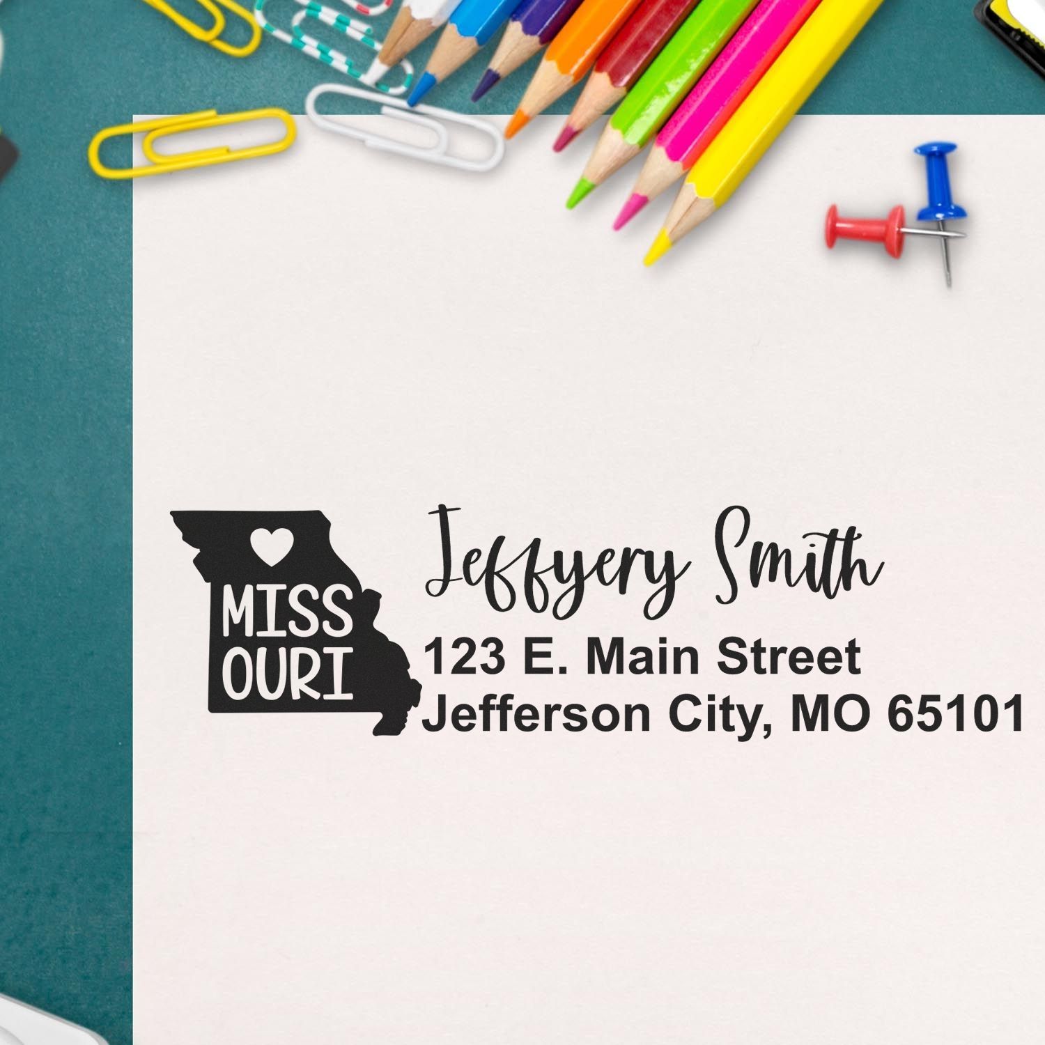 State Love of Missouri Custom Address Stamp Self-Inking on white paper, featuring a heart in Missouri's outline, surrounded by colorful pencils and paperclips on a green surface.