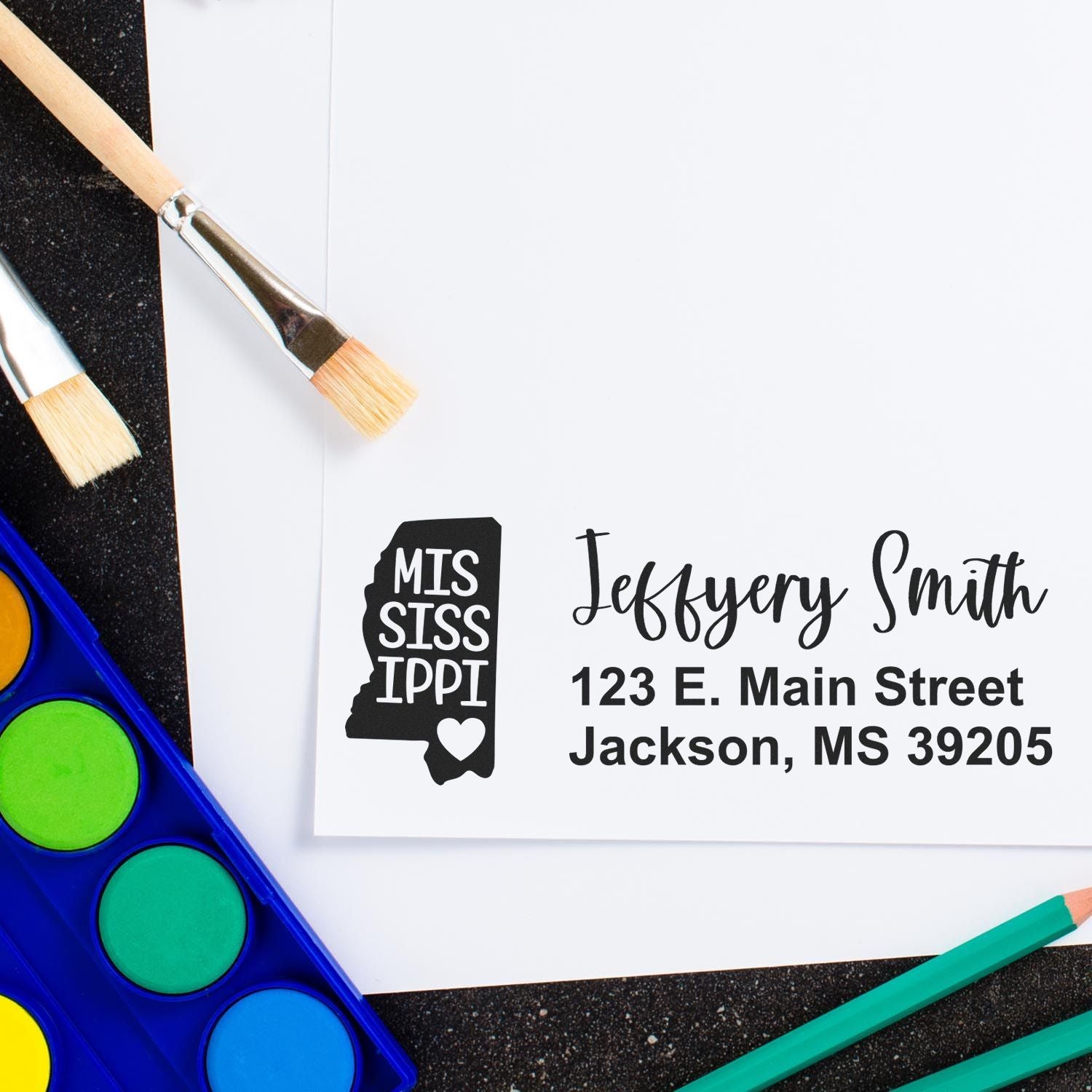 Mississippi State Love Personalized Address Stamp on white paper with brushes and colorful paints nearby, featuring a state outline and heart design with custom name and address.