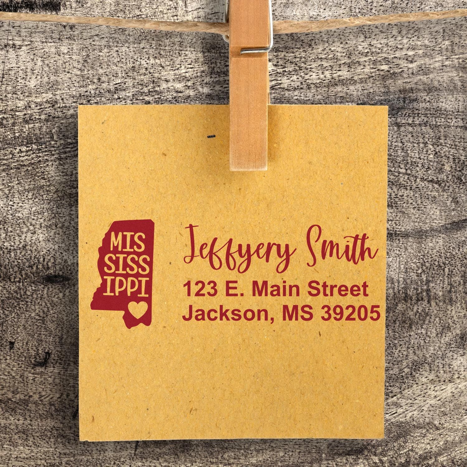 Mississippi State Love Personalized Address Stamp on brown paper, featuring a state outline and heart, with the name Jeffery Smith and address in red. Attached to a wooden surface with a clothespin.