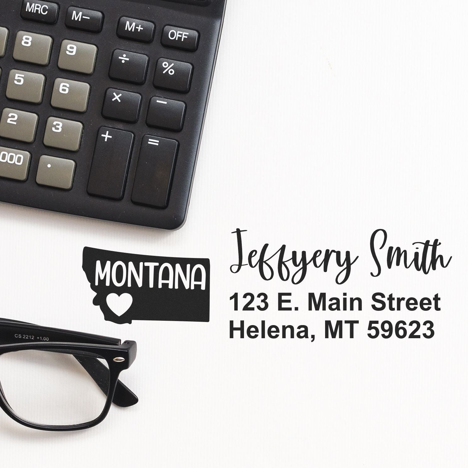 Montana State Love Personalized Address Stamp displayed on paper with a calculator and glasses nearby. The stamp shows a heart inside the state outline, with a name and address in elegant font.