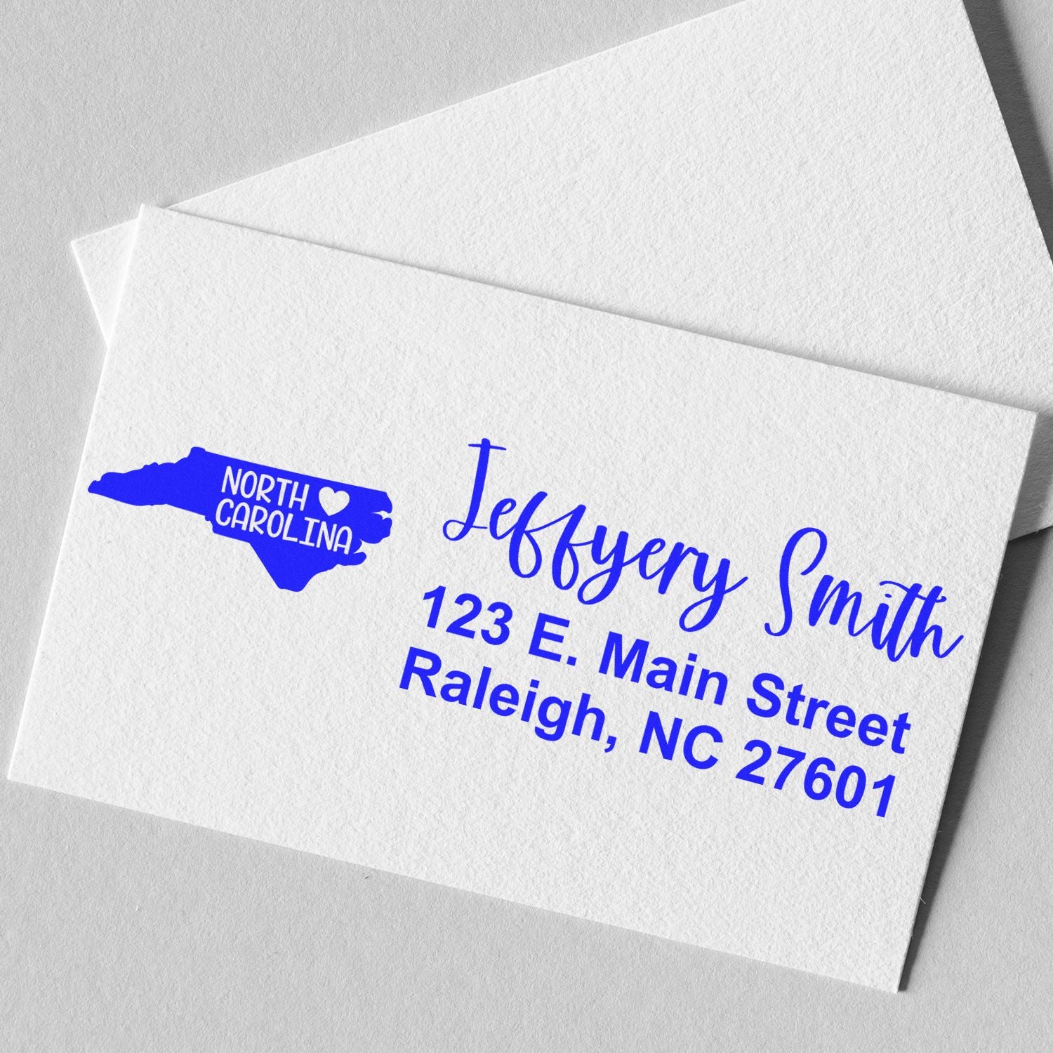 Slim North Carolina Custom Address Stamp for Envelopes displayed on a white card, featuring a blue state outline with a heart and personalized address in elegant blue font.