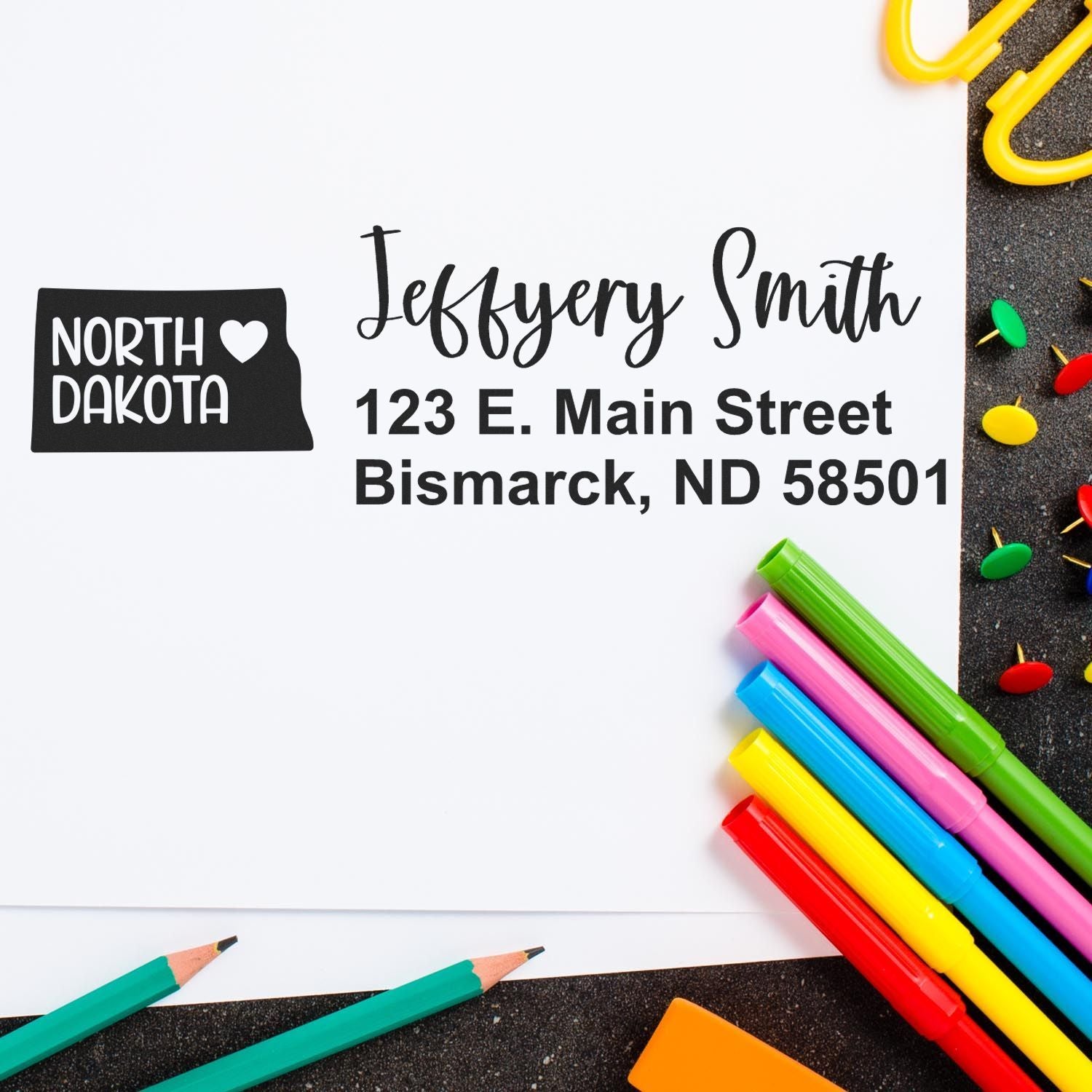 PSI Pre-Inked North Dakota State Love Customized Address Stamp on white paper with colorful pens and paperclips, featuring a heart design and personalized address for Jeffery Smith in Bismarck, ND.