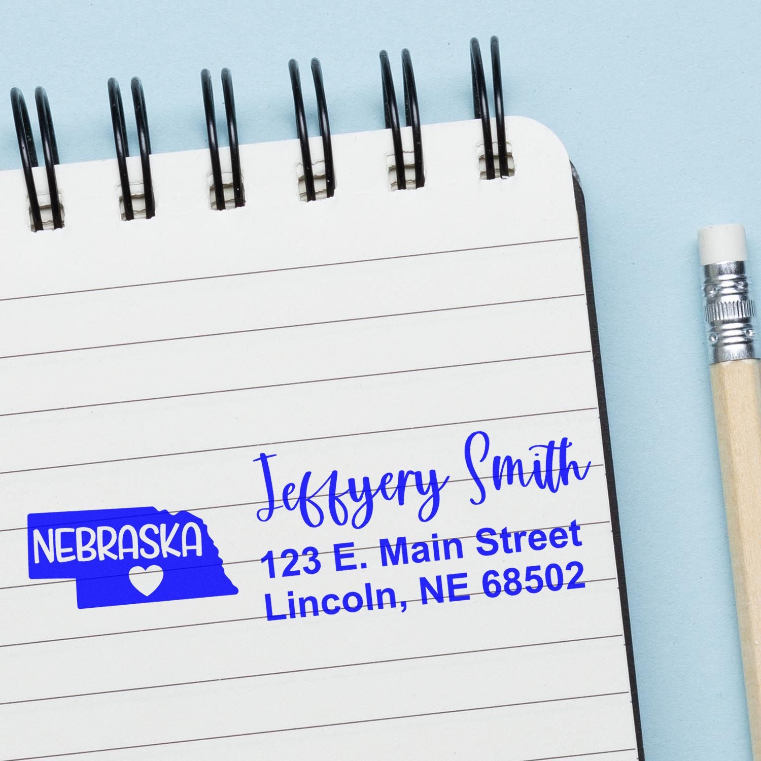 Nebraska State Love Personalized Address Stamp on a notebook page, featuring a blue Nebraska state outline with a heart, and sample address text in elegant font. Pencil placed beside the notebook.