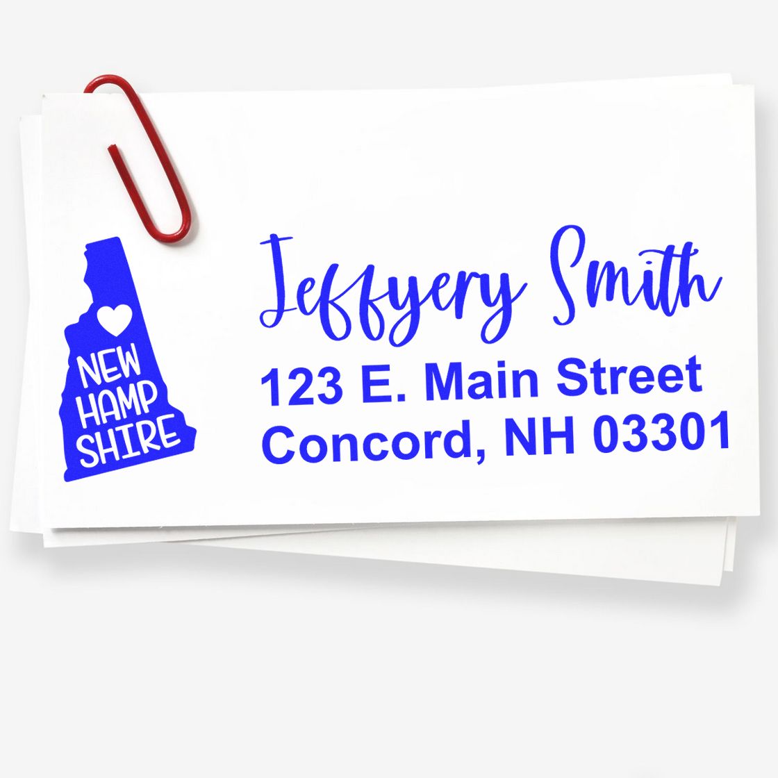 Slim New Hampshire Custom Address Stamp for Envelopes in blue ink on white paper, featuring a state outline with a heart. Includes name, street, city, and zip code. Red paperclip attached.