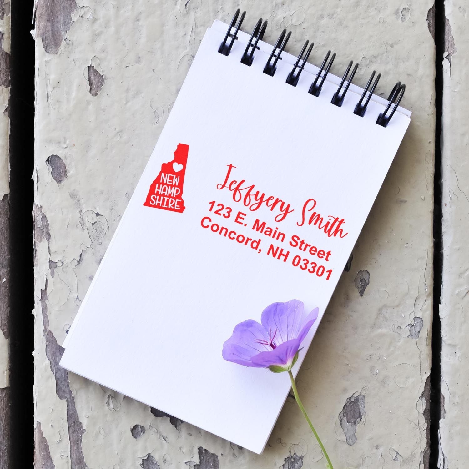 A notepad on a rustic surface features the State Love of New Hampshire Custom Address Stamp Self-Inking in red, displaying a name and address with a purple flower nearby.