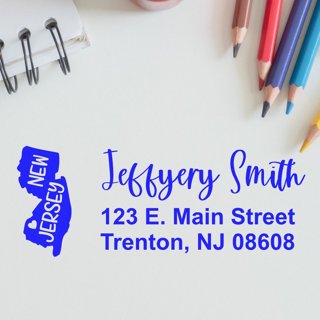 Slim New Jersey Custom Address Stamp for Envelopes displayed on a white surface with colorful pencils and a notebook, featuring a blue map of New Jersey and sample address text in elegant font.