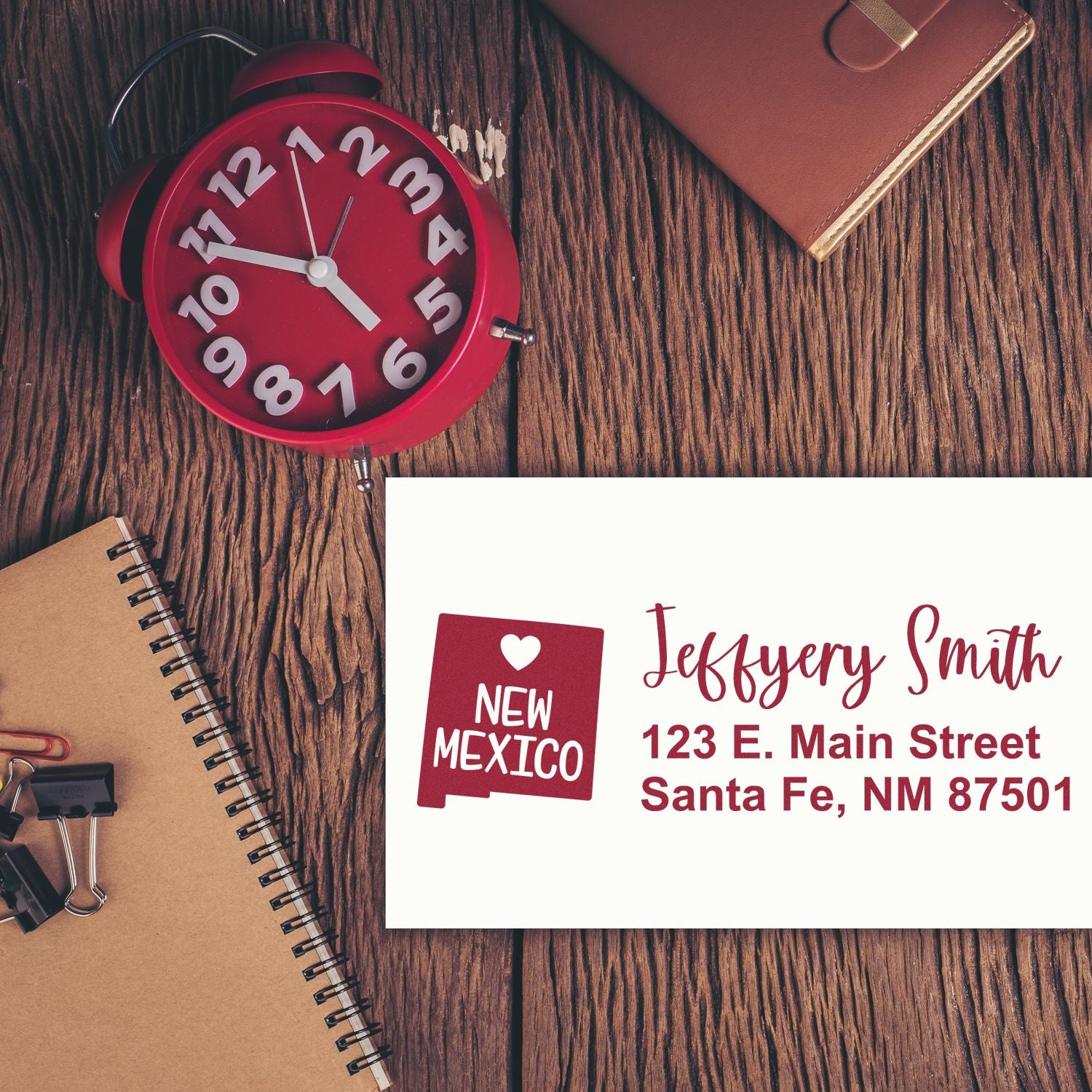 New Mexico State Love Personalized Address Stamp on envelope with red clock, notebook, and leather items on wooden table. Stamp shows name and address in red with state outline and heart.