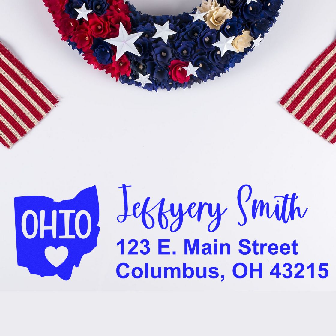 State Love of Ohio Custom Address Stamp Self-Inking displayed with a patriotic wreath and striped fabric, featuring a sample address: Jeffery Smith, 123 E. Main Street, Columbus, OH 43215.