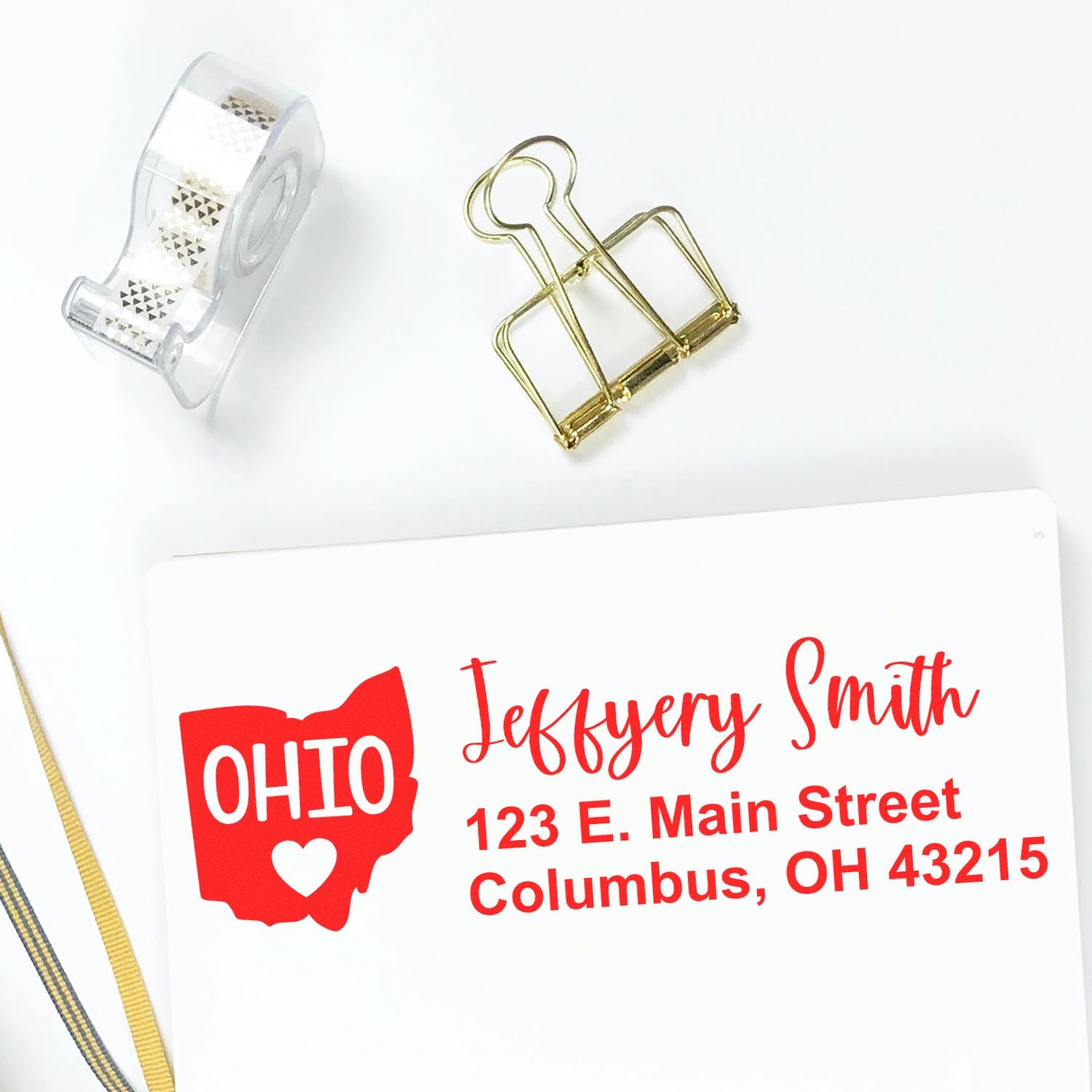 State Love of Ohio Custom Address Stamp Self-Inking displayed on white paper with red text, featuring an Ohio map design. Nearby are a tape dispenser, gold clip, and ribbon.