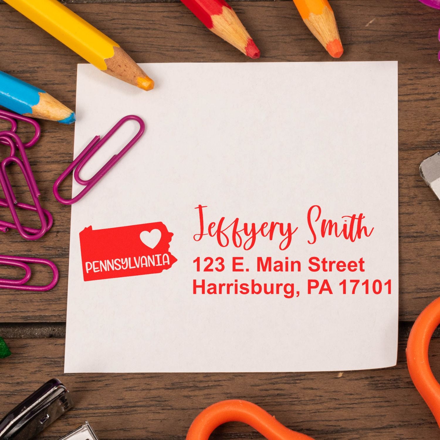 State Love of Pennsylvania Custom Address Stamp Self-Inking on a white card with red text, surrounded by colorful pencils and paperclips on a wooden surface.