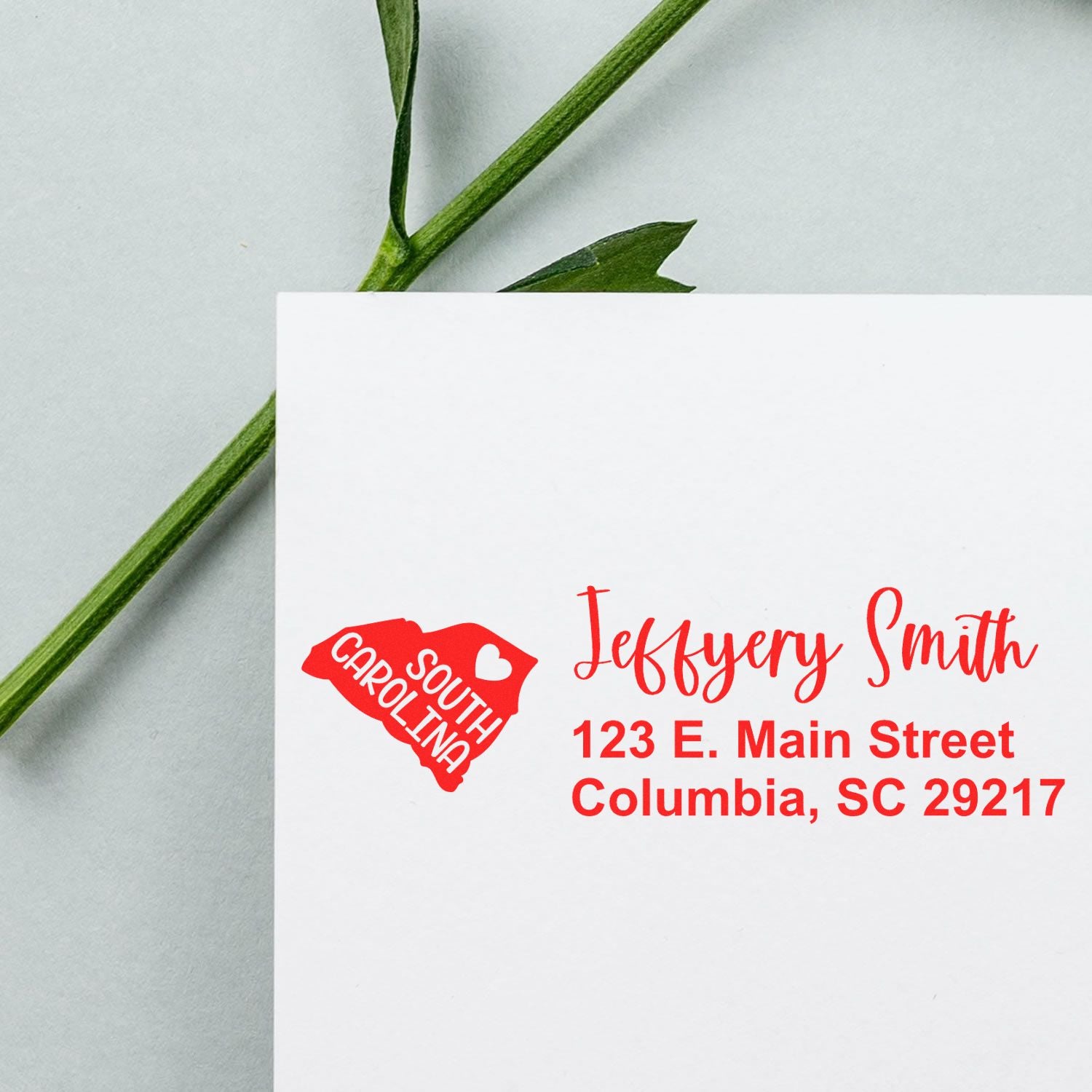 Envelope with South Carolina State Love Personalized Address Stamp in red, featuring a heart and state outline, next to a green stem on a light background.