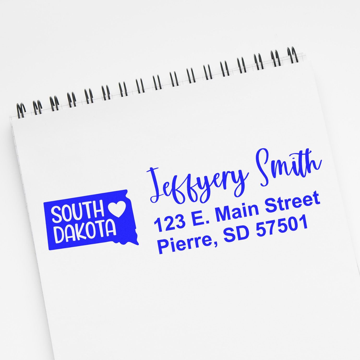 State Love of South Dakota Custom Address Stamp Self-Inking displayed on a notepad with the text Jeffery Smith, 123 E. Main Street, Pierre, SD 57501 in blue ink.