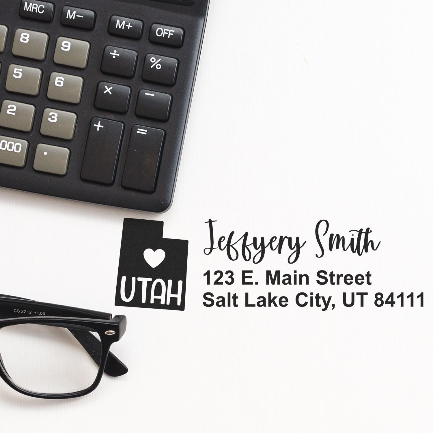 Utah State Love Personalized Address Stamp next to a calculator and glasses on a white surface, featuring a heart and 'UTAH' design with sample address: Jeffery Smith, 123 E. Main Street, Salt Lake City, UT 84111.