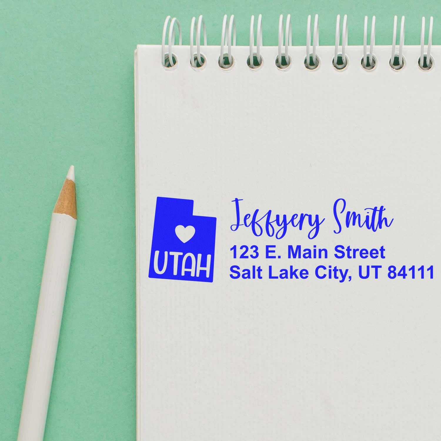 State Love of Utah Custom Address Stamp Self-Inking on a notepad. Blue ink shows a Utah state outline with a heart, personalized with name and address. Pencil nearby on a green background.