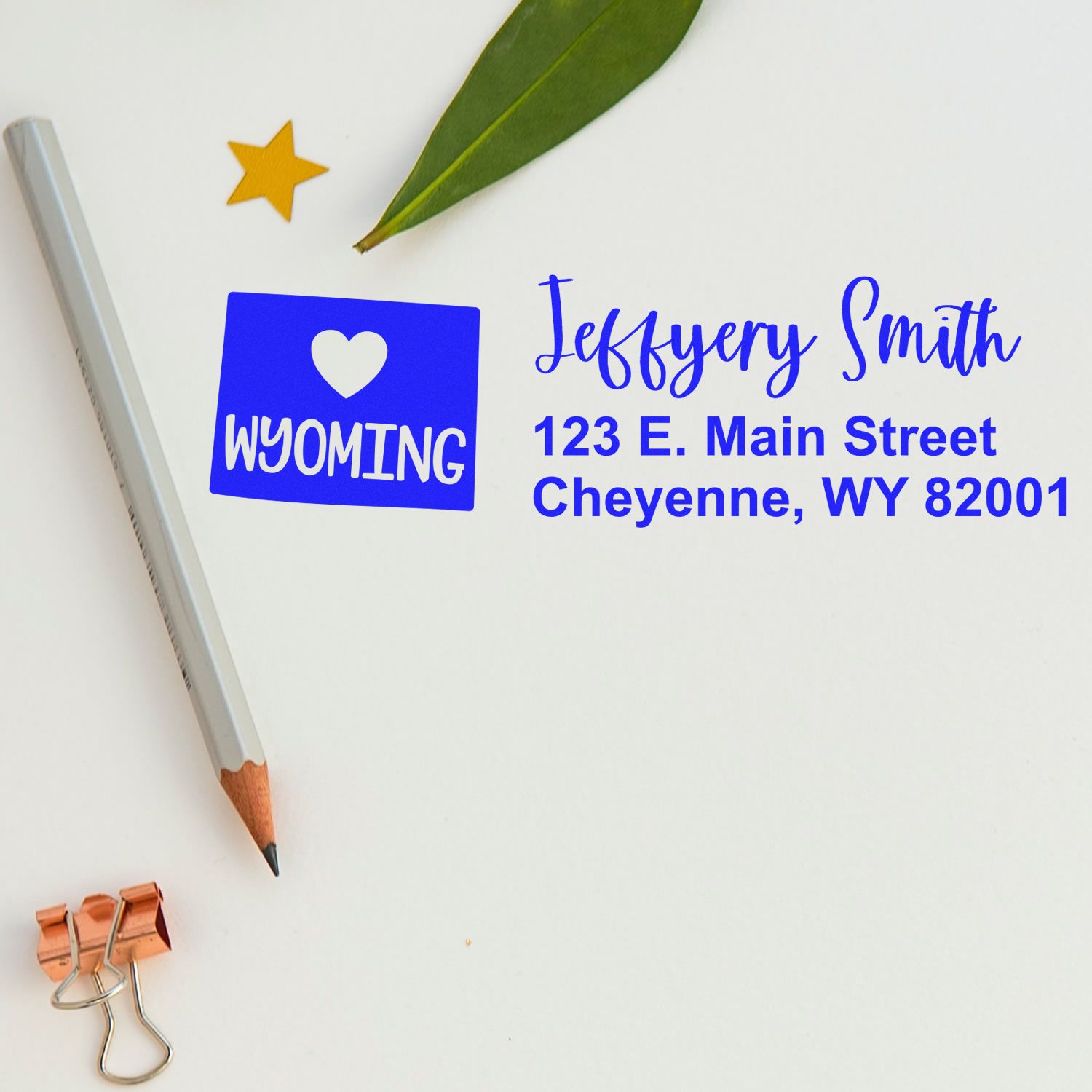 Slim Wyoming Custom Address Stamp for Envelopes displayed on a white surface with a pencil, leaf, and star sticker. The stamp shows a heart, Wyoming, and an address in blue text.