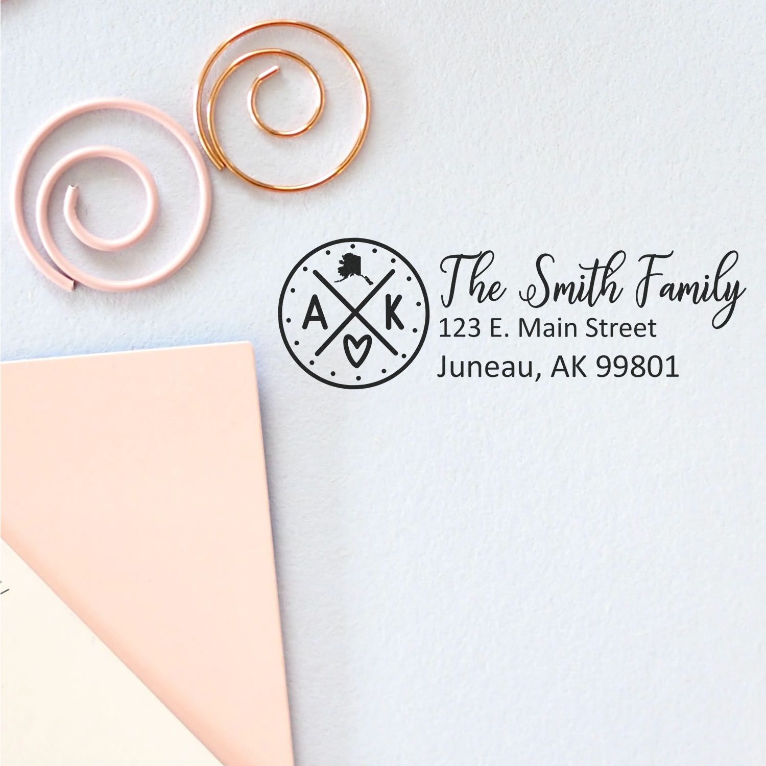 Slim Alaska Customized Pre-Inked Address Stamp on light surface with pink and gold spirals, featuring 'The Smith Family, 123 E. Main Street, Juneau, AK 99801' in elegant black font.