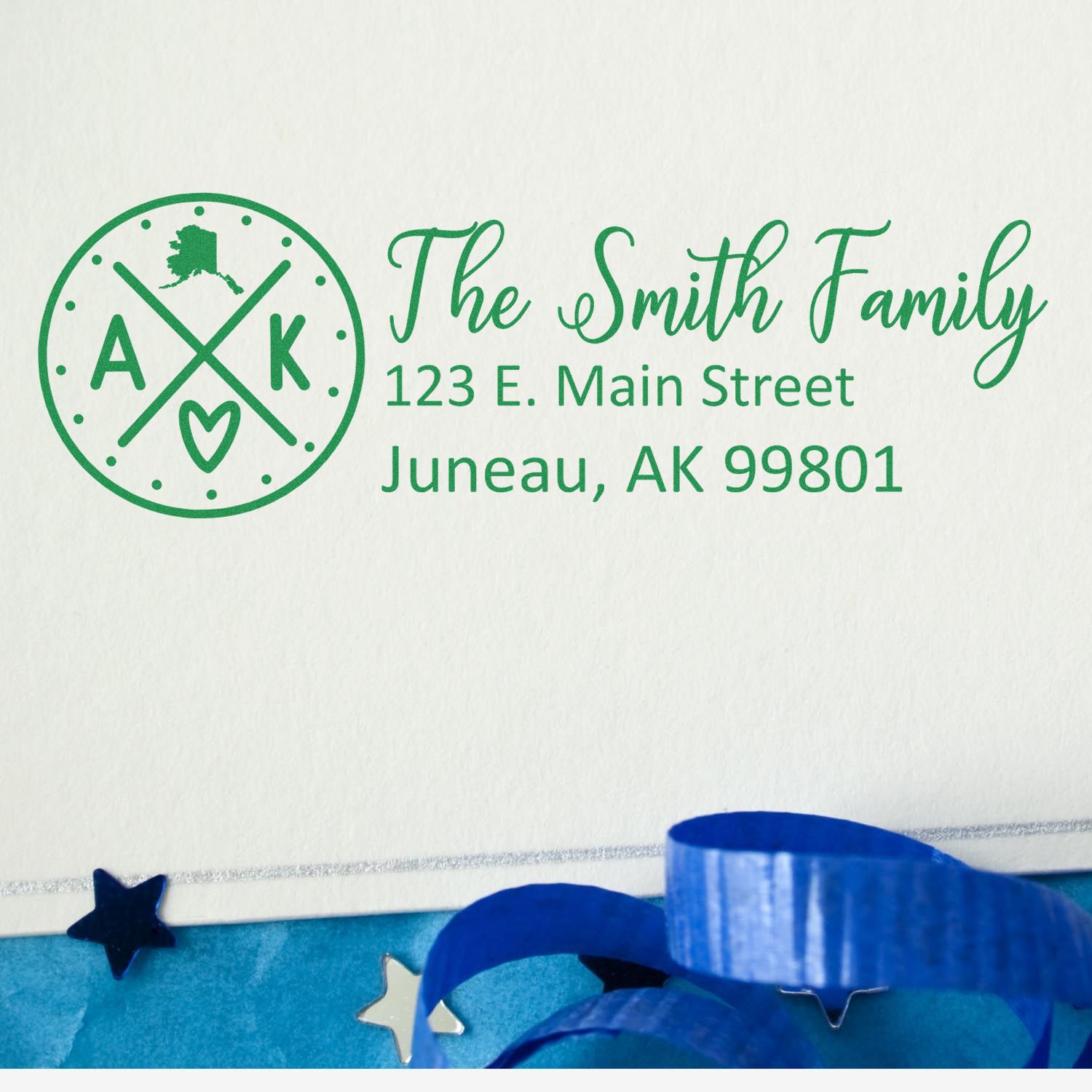 Self-Inking State Love of Alaska Custom Address Stamp on an envelope with The Smith Family, 123 E. Main Street, Juneau, AK 99801 in green ink, surrounded by blue ribbon and star confetti.