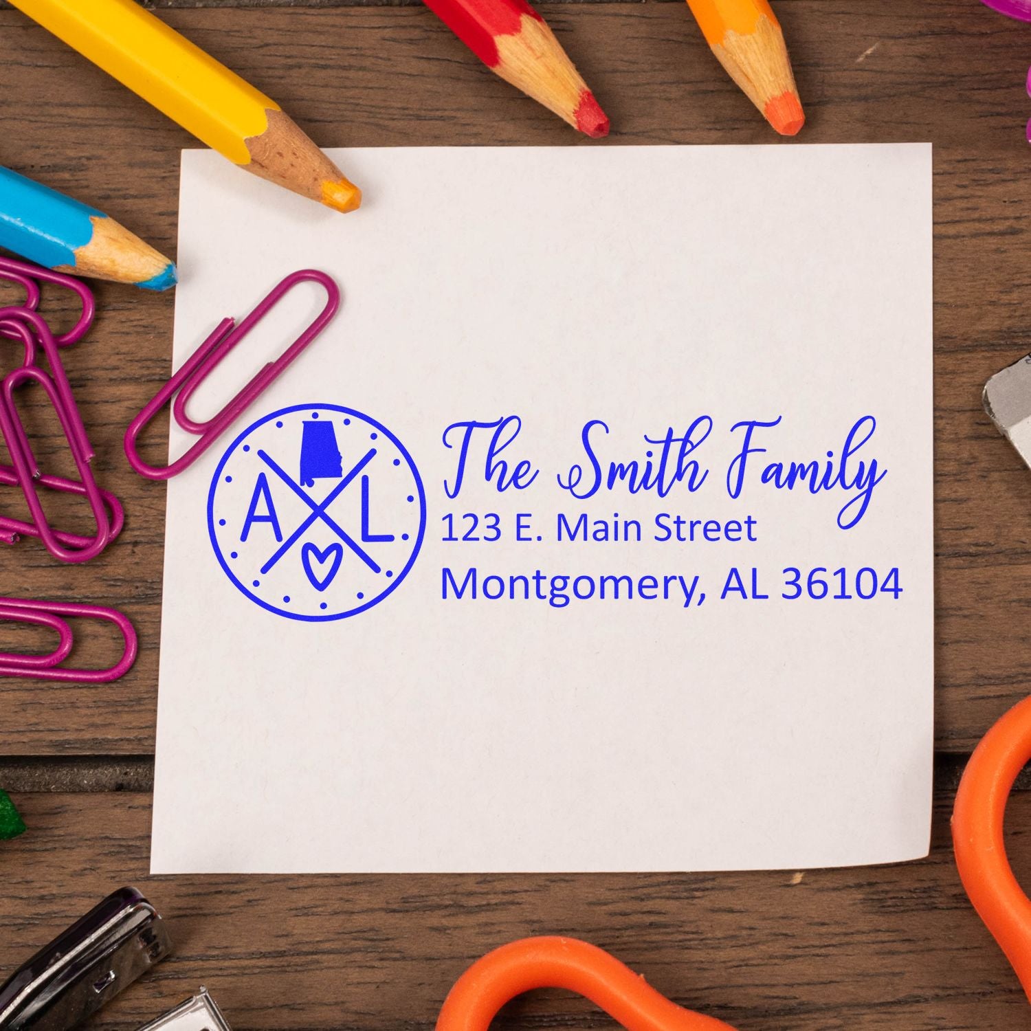 Alabama State Pride Customized Address Stamp on paper with The Smith Family and address, surrounded by colorful pencils and paperclips on a wooden surface.