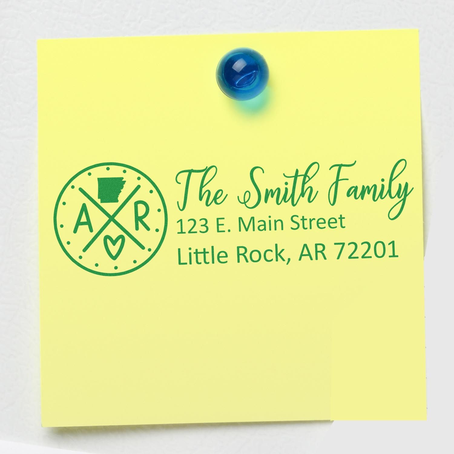 Yellow note with green ink showing the Self-Inking State Love of Arkansas Custom Address Stamp design, featuring The Smith Family and an address in Little Rock, AR, with a blue magnet on top.