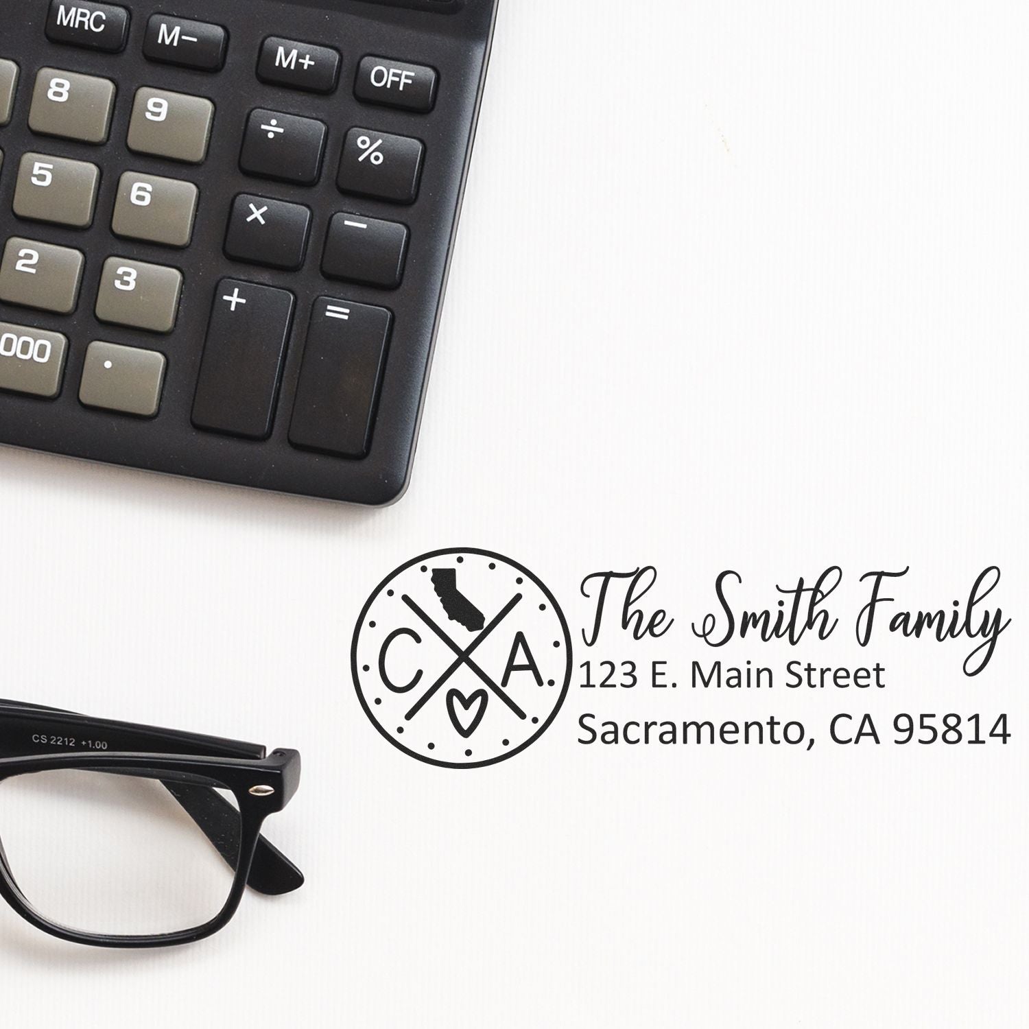 A Self-Inking State Love of California Custom Address Stamp on a desk with glasses and a calculator, featuring a personalized address for the Smith Family in Sacramento, CA.