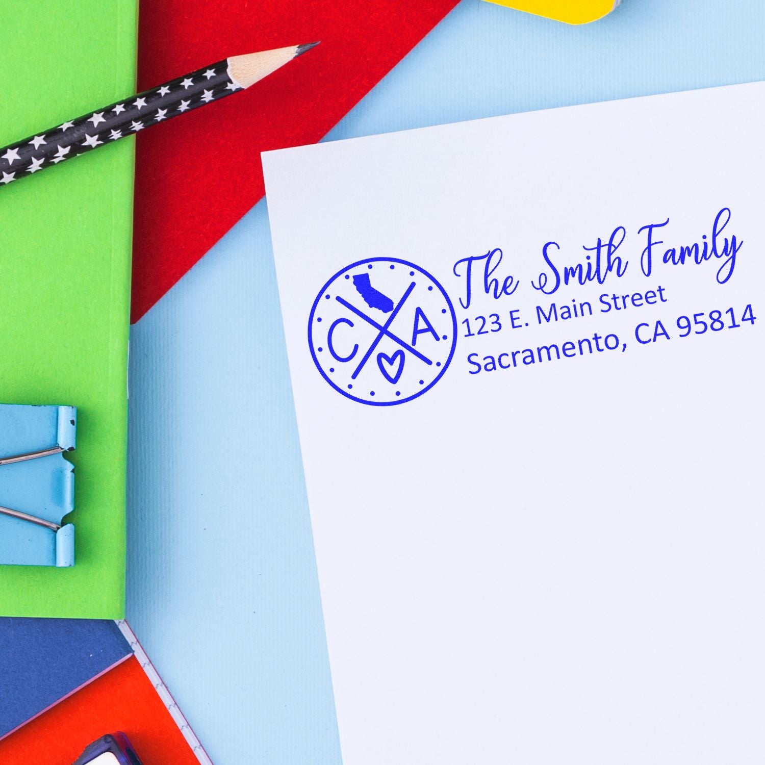 A colorful desk with paper and clips features a stamped envelope using the Self-Inking State Love of California Custom Address Stamp, displaying a personalized address in blue ink.