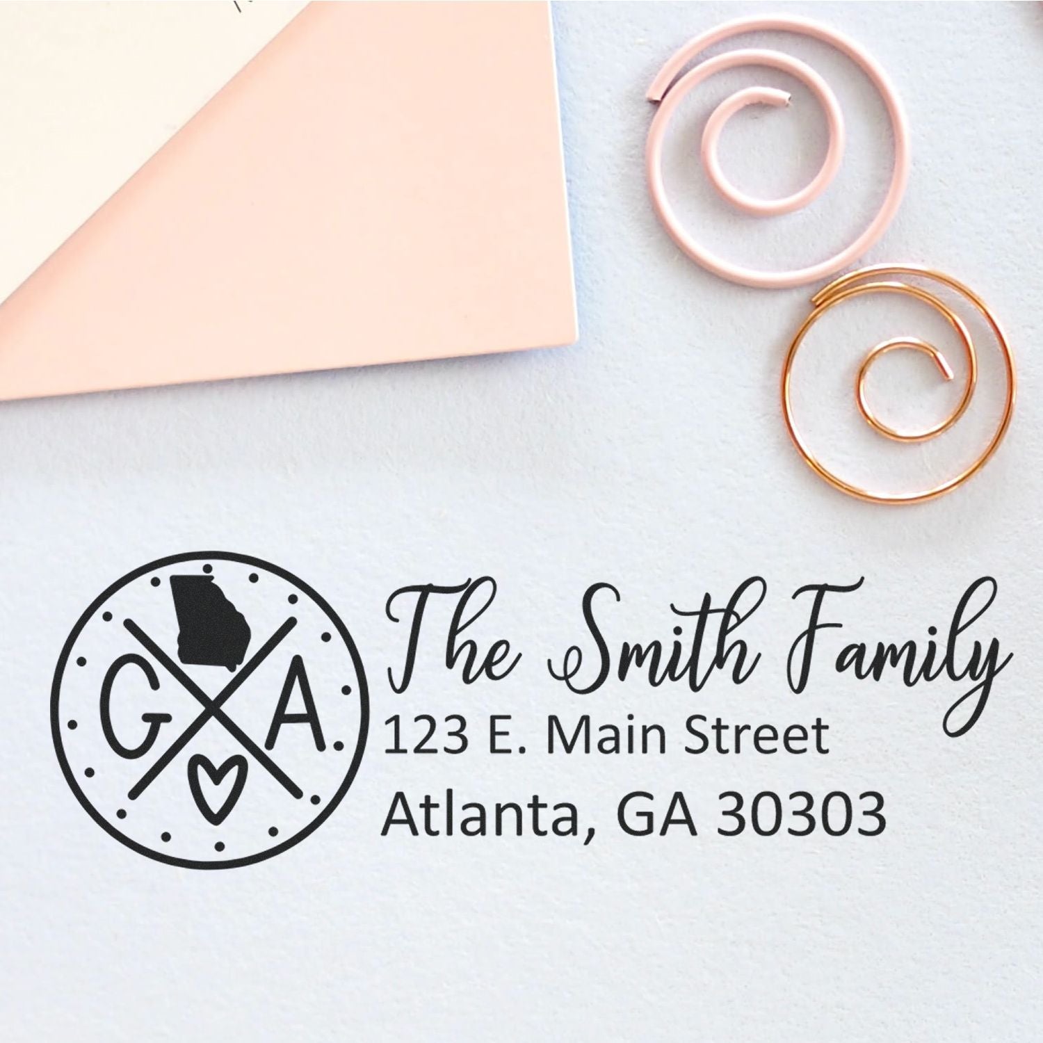 Slim Georgia Customized Pre-Inked Address Stamp on paper with 'The Smith Family, 123 E. Main Street, Atlanta, GA 30303' design. Includes decorative paper clips and envelope on a light background.