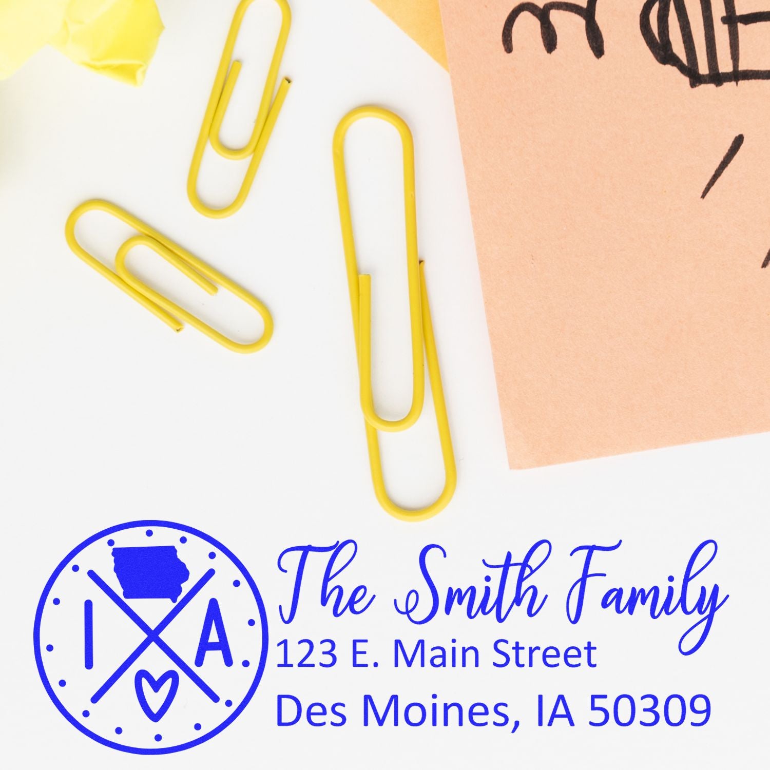 Image of the Iowa State Pride Customized Address Stamp with The Smith Family and an Iowa map design, placed on a desk with yellow paperclips and a peach-colored paper.