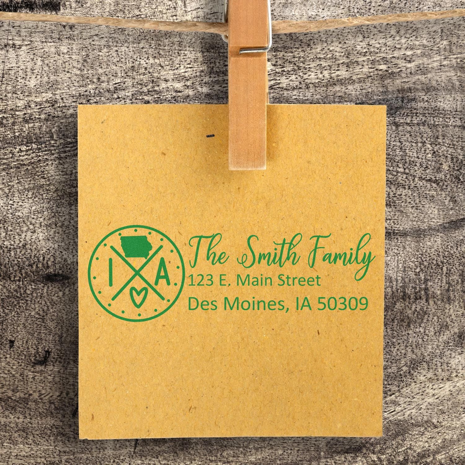 A brown card with green ink shows The Smith Family, 123 E. Main Street, Des Moines, IA 50309 using the Slim Iowa Customized Pre-Inked Address Stamp, clipped to a rustic wooden background.