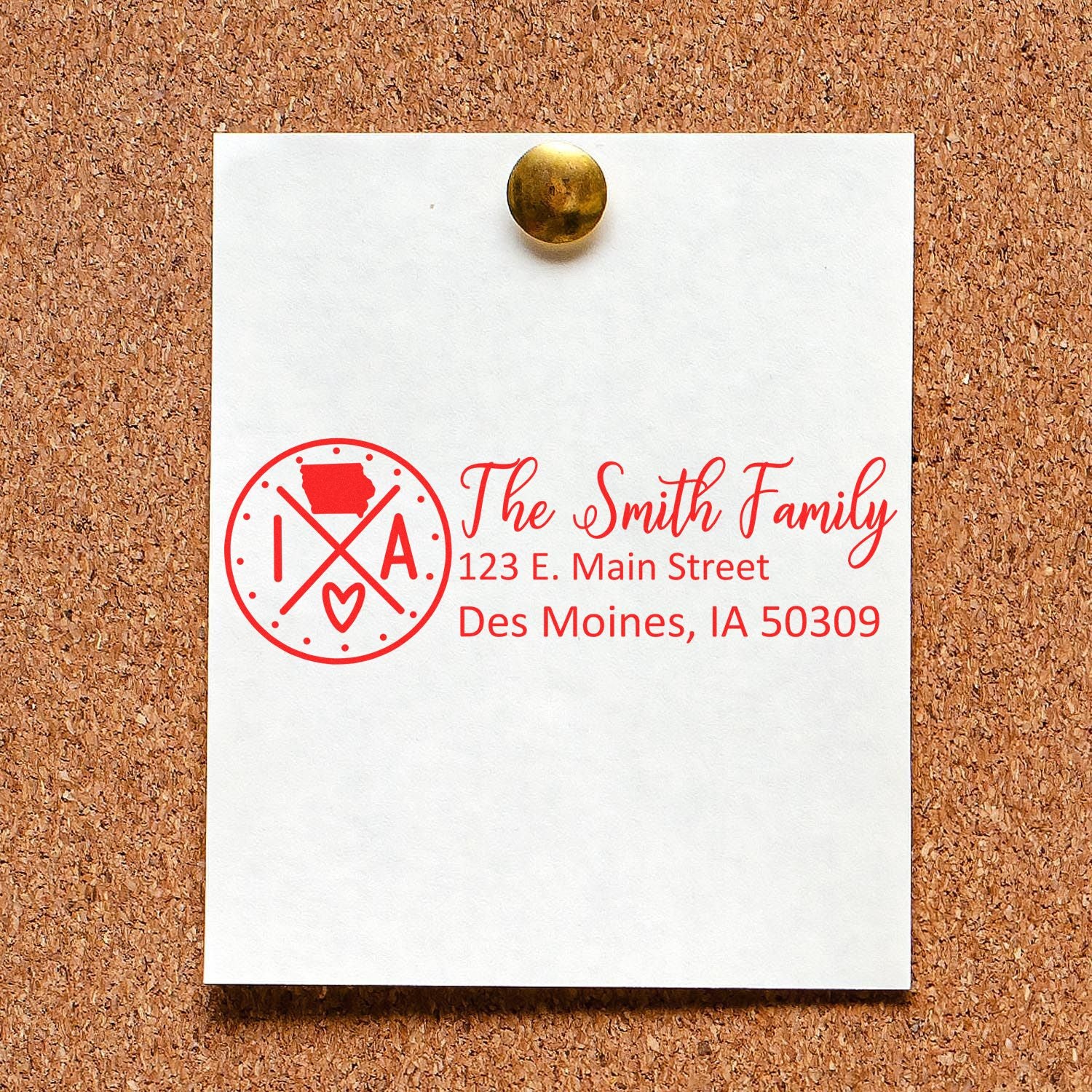 PSI Pre-Inked Iowa State Pride Customized Address Stamp on a corkboard, displaying The Smith Family, 123 E. Main Street, Des Moines, IA 50309 in red ink with an Iowa-themed design.