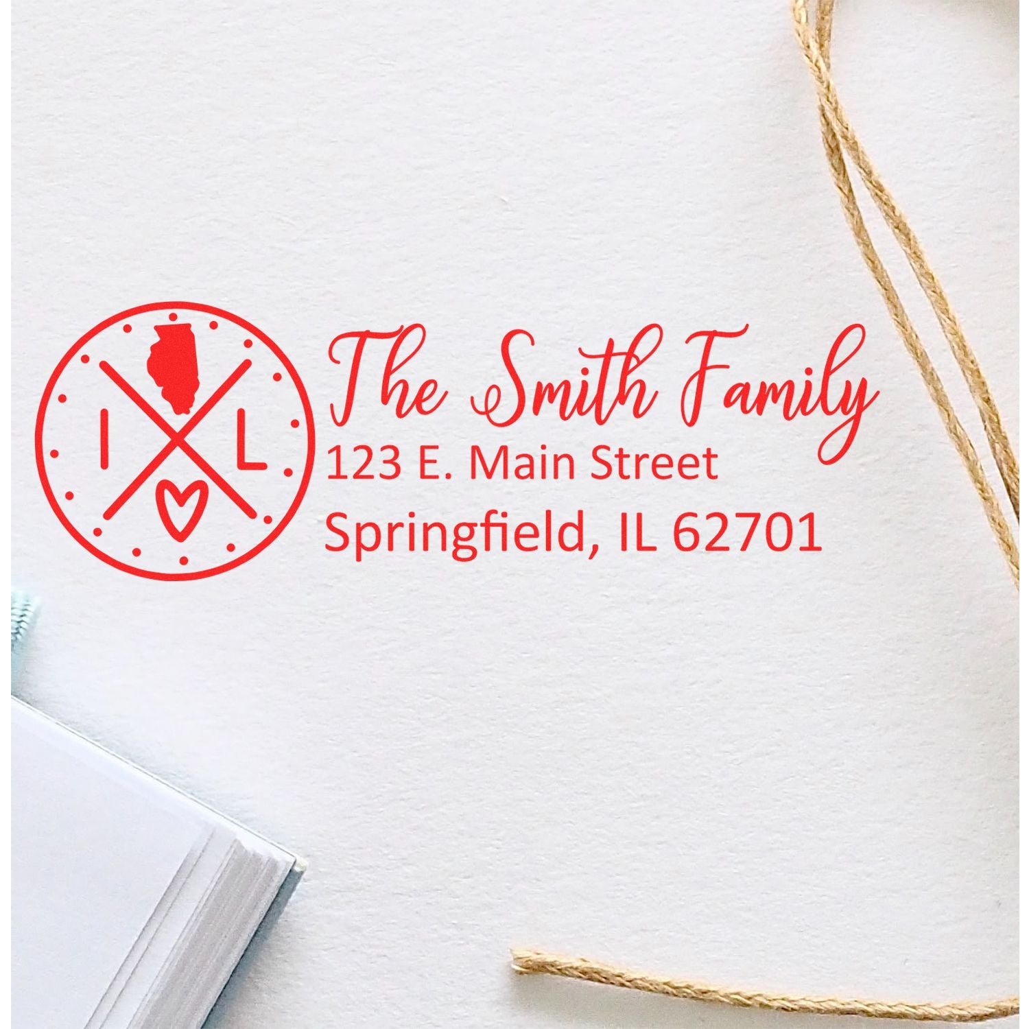 Illinois State Pride Customized Address Stamp with red design featuring state outline, family name, and address on white paper. Nearby are twine and a notebook, creating a personalized stationery look.