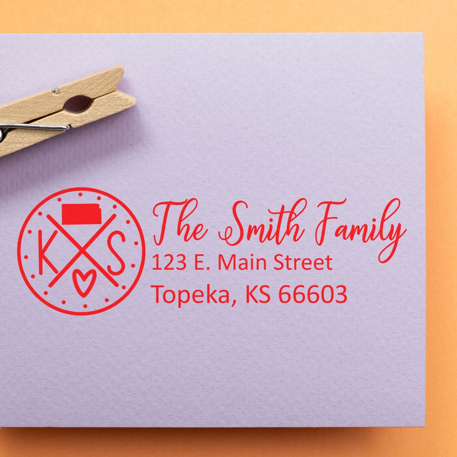 Kansas State Pride Customized Address Stamp on a lavender envelope with red ink, displaying The Smith Family, 123 E. Main Street, Topeka, KS 66603. A wooden clothespin is clipped to the envelope.