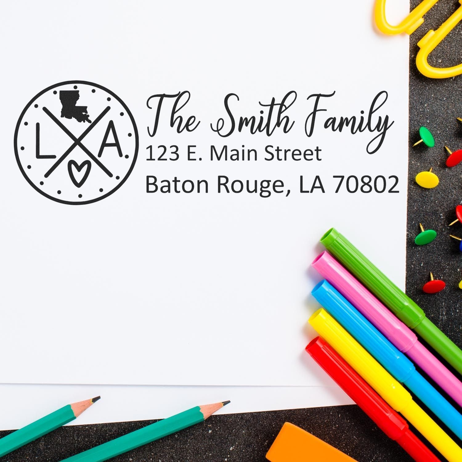 A Slim Louisiana Customized Pre-Inked Address Stamp marks an envelope with The Smith Family, 123 E. Main Street, Baton Rouge, LA 70802 beside colorful pens and paperclips on a dark surface.