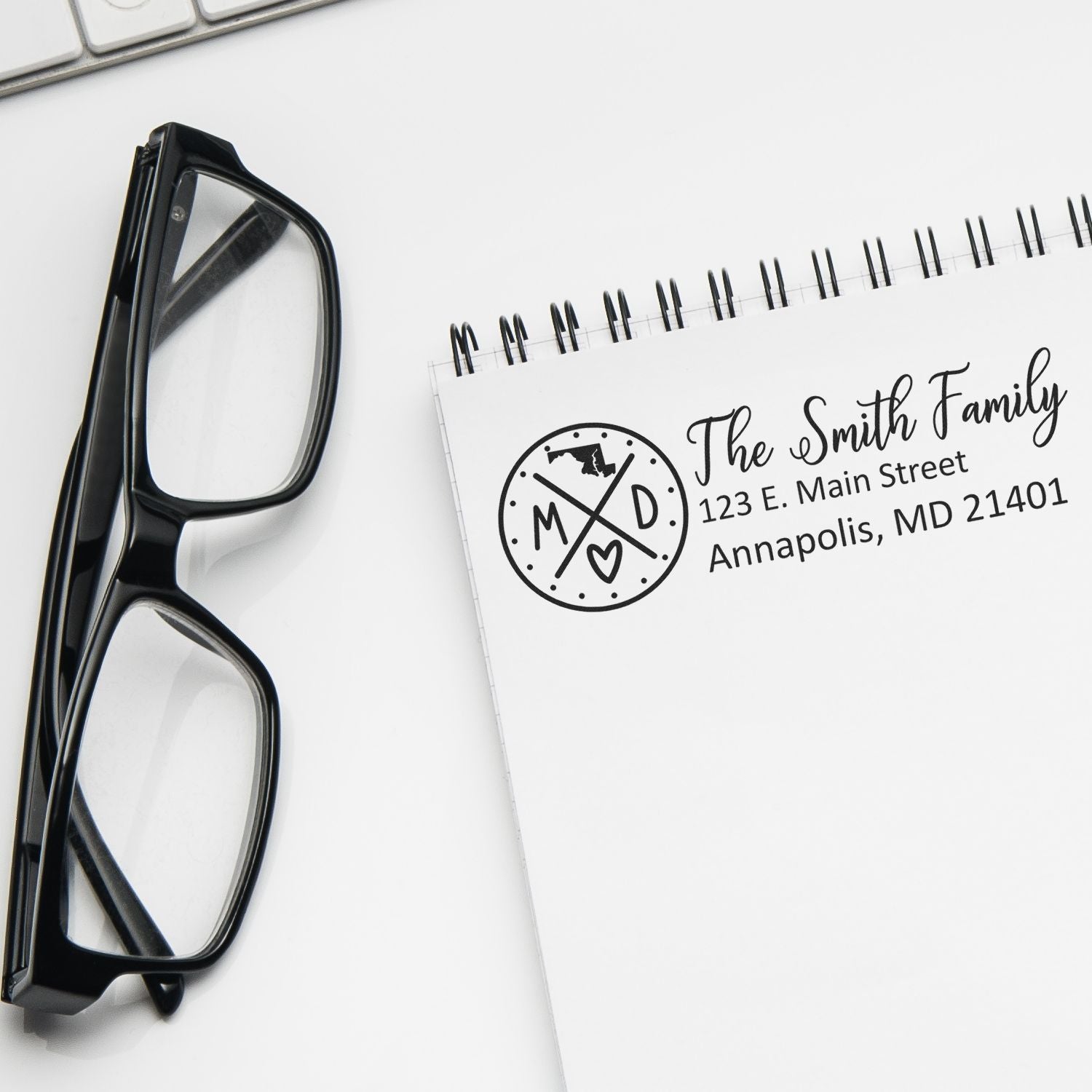 PSI Pre-Inked Maryland State Pride Customized Address Stamp on a notepad with glasses nearby, displaying The Smith Family, 123 E. Main Street, Annapolis, MD 21401 in elegant font.