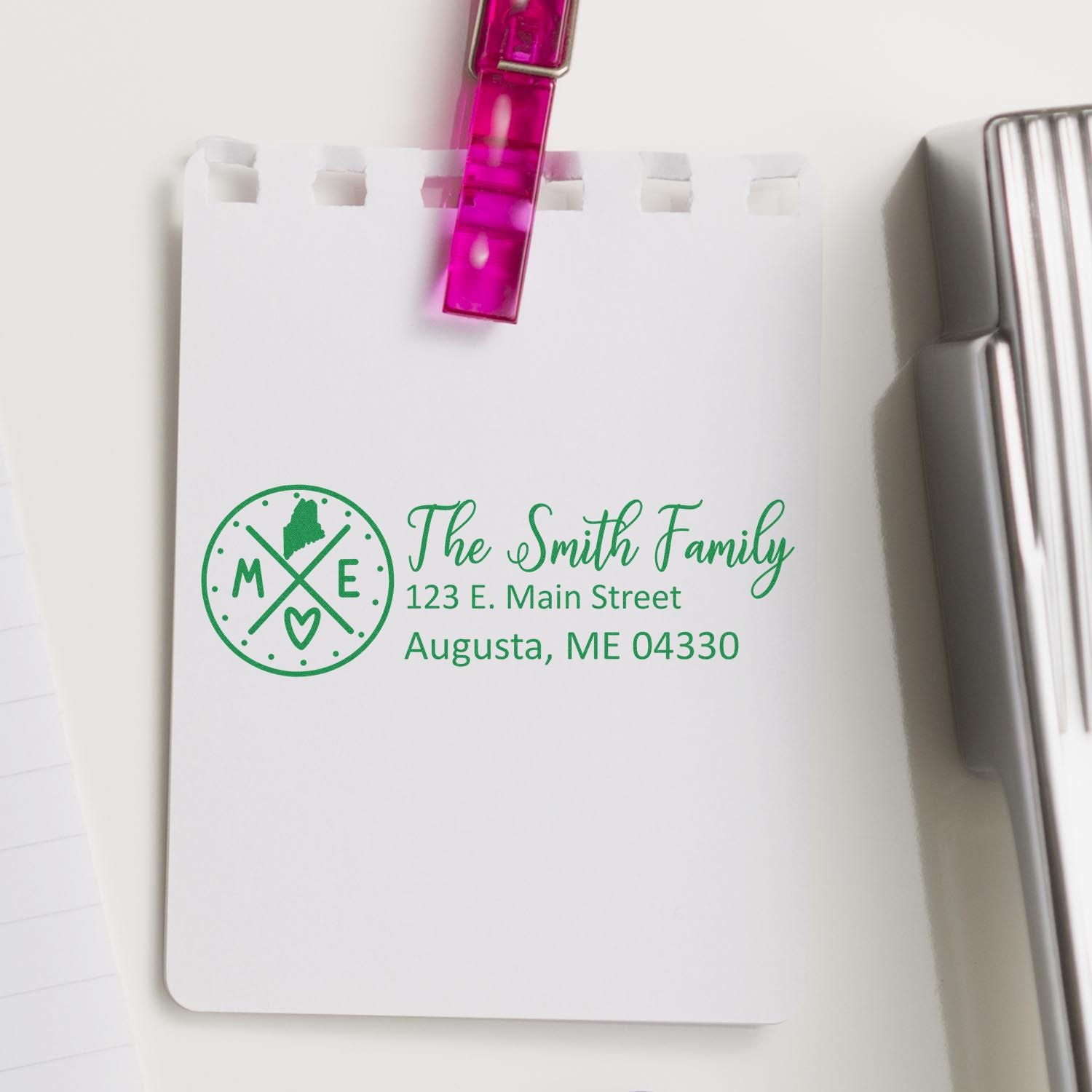 Maine State Pride Customized Address Stamp on a notepad, featuring The Smith Family and an Augusta, ME address in green ink, with a pink clip and a stapler nearby.