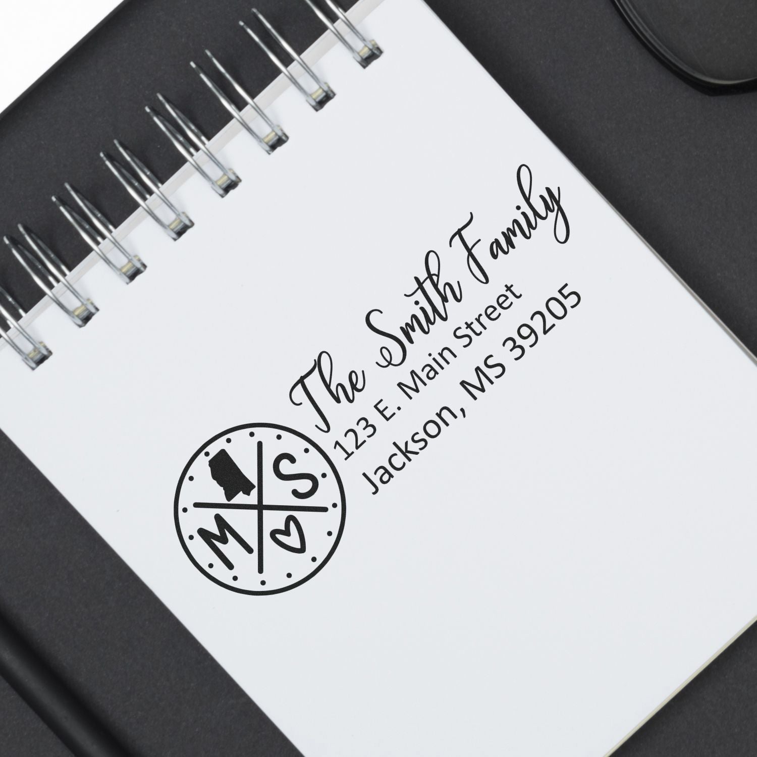 Slim Mississippi Customized Pre-Inked Address Stamp on a notebook, displaying The Smith Family, 123 E. Main Street, Jackson, MS 39205 with a circular MS design. Black ink on white paper.