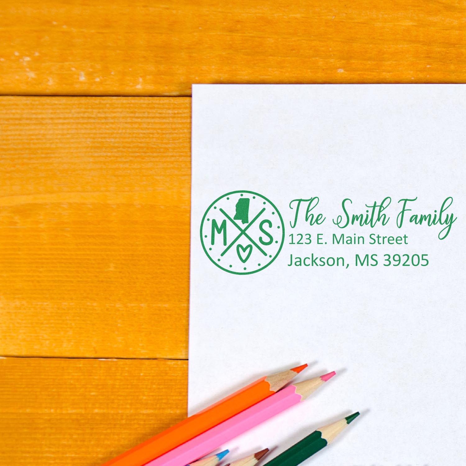 Self-Inking State Love of Mississippi Custom Address Stamp on white paper with green ink, featuring a heart and state outline. Colored pencils nearby on a wooden surface.