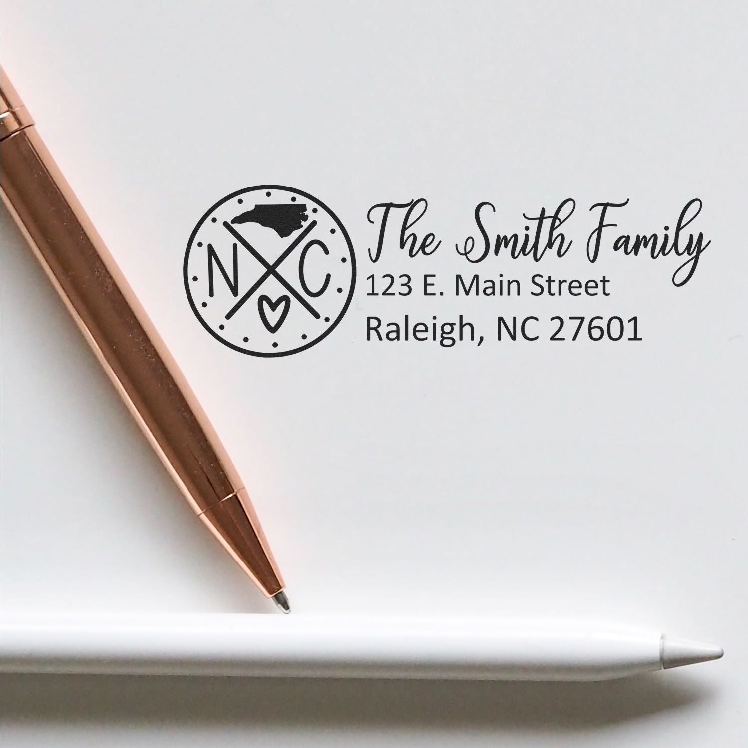 PSI Pre-Inked North Carolina State Pride Customized Address Stamp on white paper with a rose gold pen and white pencil. Stamp shows NC with a heart and address for The Smith Family in Raleigh.