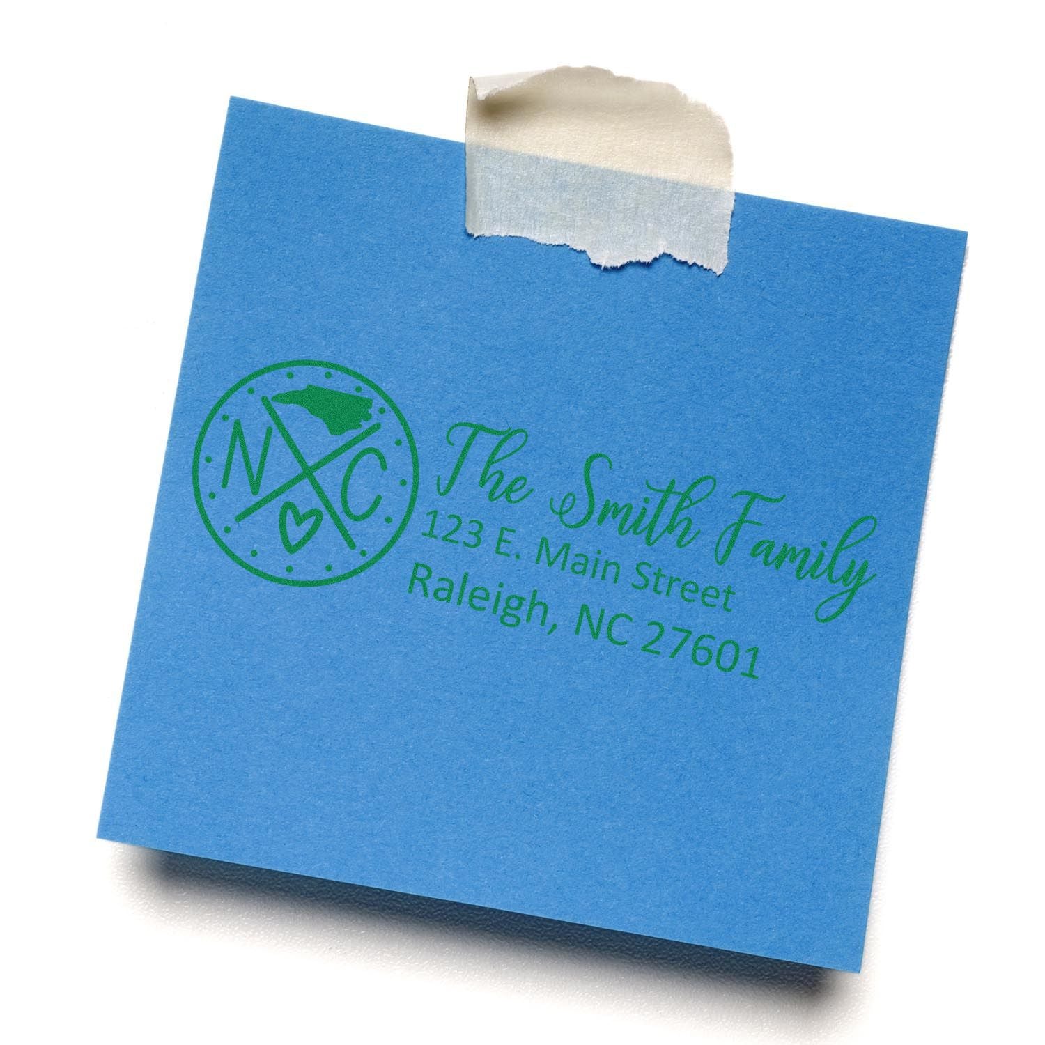 Blue paper with a green imprint from the PSI Pre-Inked North Carolina State Pride Customized Address Stamp, featuring The Smith Family and an address in Raleigh, NC, with a state outline and heart.