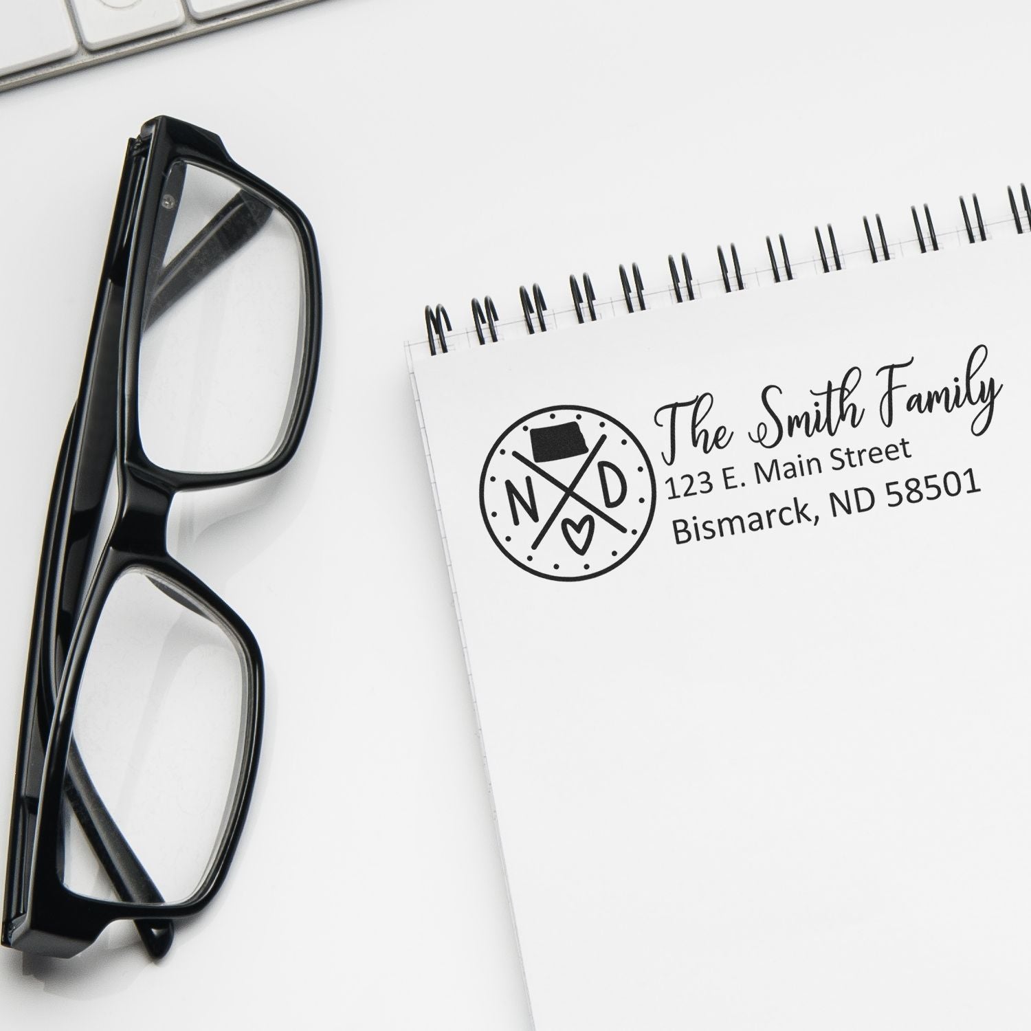 A PSI Pre-Inked North Dakota State Pride Customized Address Stamp on a notepad, next to black glasses and a keyboard, displaying The Smith Family with an ND state design.