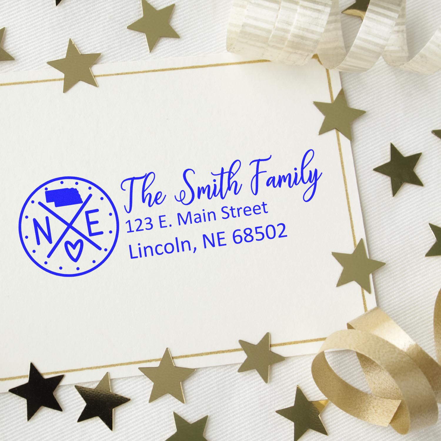 Slim Nebraska Customized Pre-Inked Address Stamp on a white envelope with gold stars and ribbon, displaying The Smith Family, 123 E. Main Street, Lincoln, NE 68502 in blue ink.