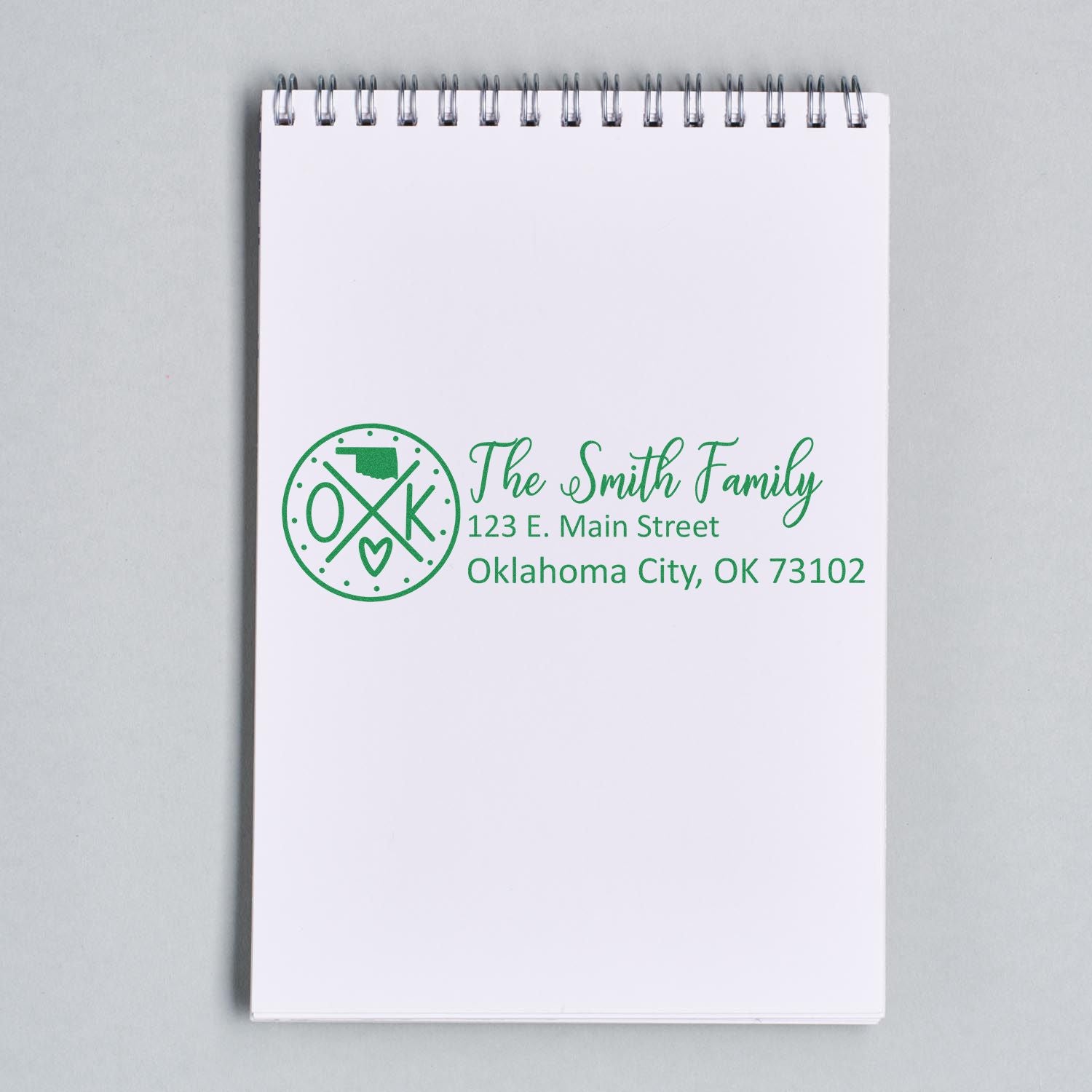 Slim Oklahoma Customized Pre-Inked Address Stamp on a notepad, displaying The Smith Family, 123 E. Main Street, Oklahoma City, OK 73102 in green ink with a circular design featuring OK and a heart.